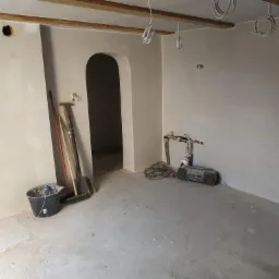 Wnętrze w trakcie prac wykończeniowych: ściany pokryte świeżą warstwą gładzi, widoczne narzędzia malarskie, wiązki kabli zwisające z drewnianego stropu i otwór w ścianie czekający na osprzęt...