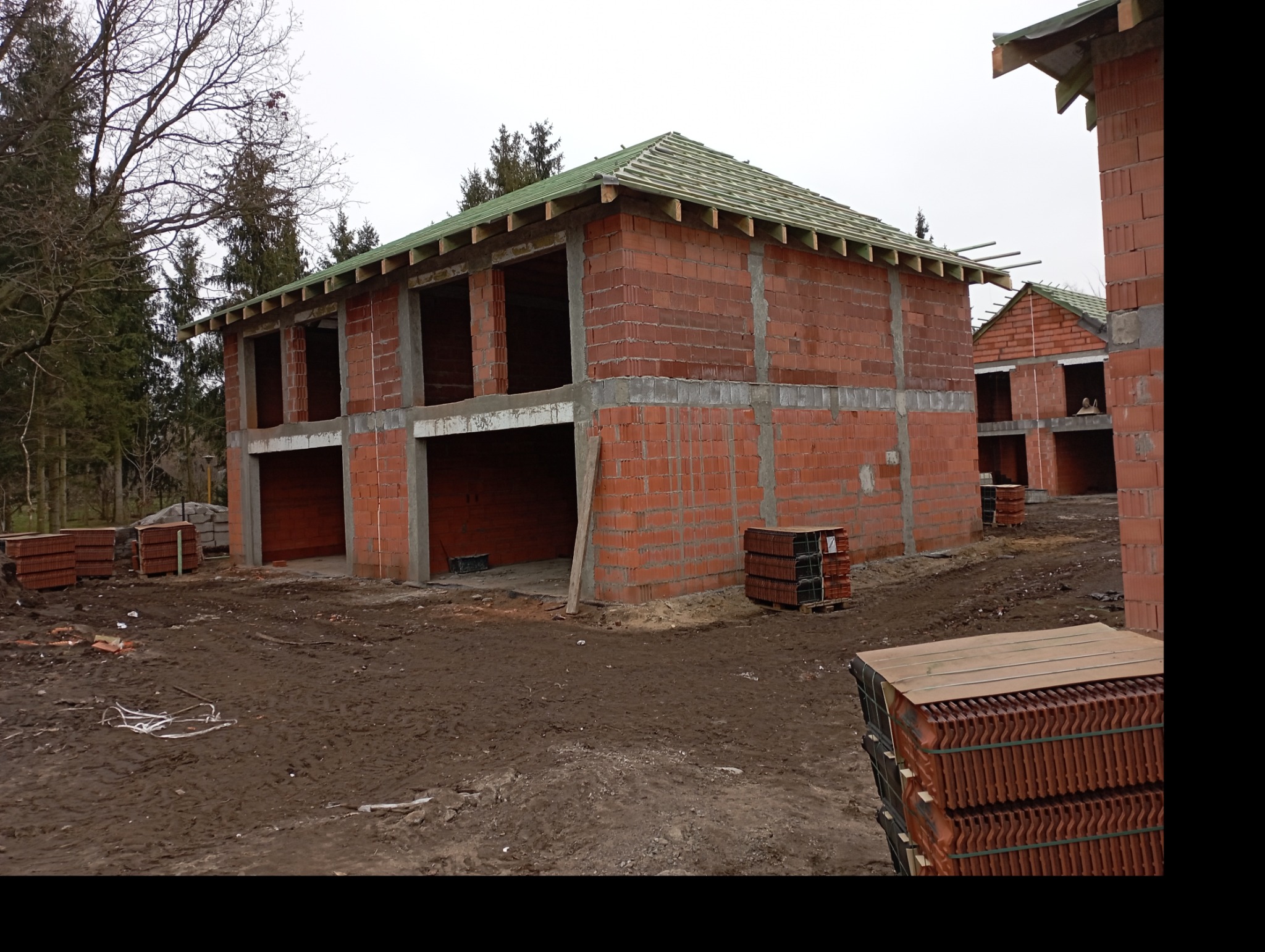 Wznoszenie dwukondygnacyjnego domu z czerwonej cegły ceramicznej, widoczne otwory okienne i garażowe, przygotowane palety z dachówką na placu budowy, w tle drzewa.