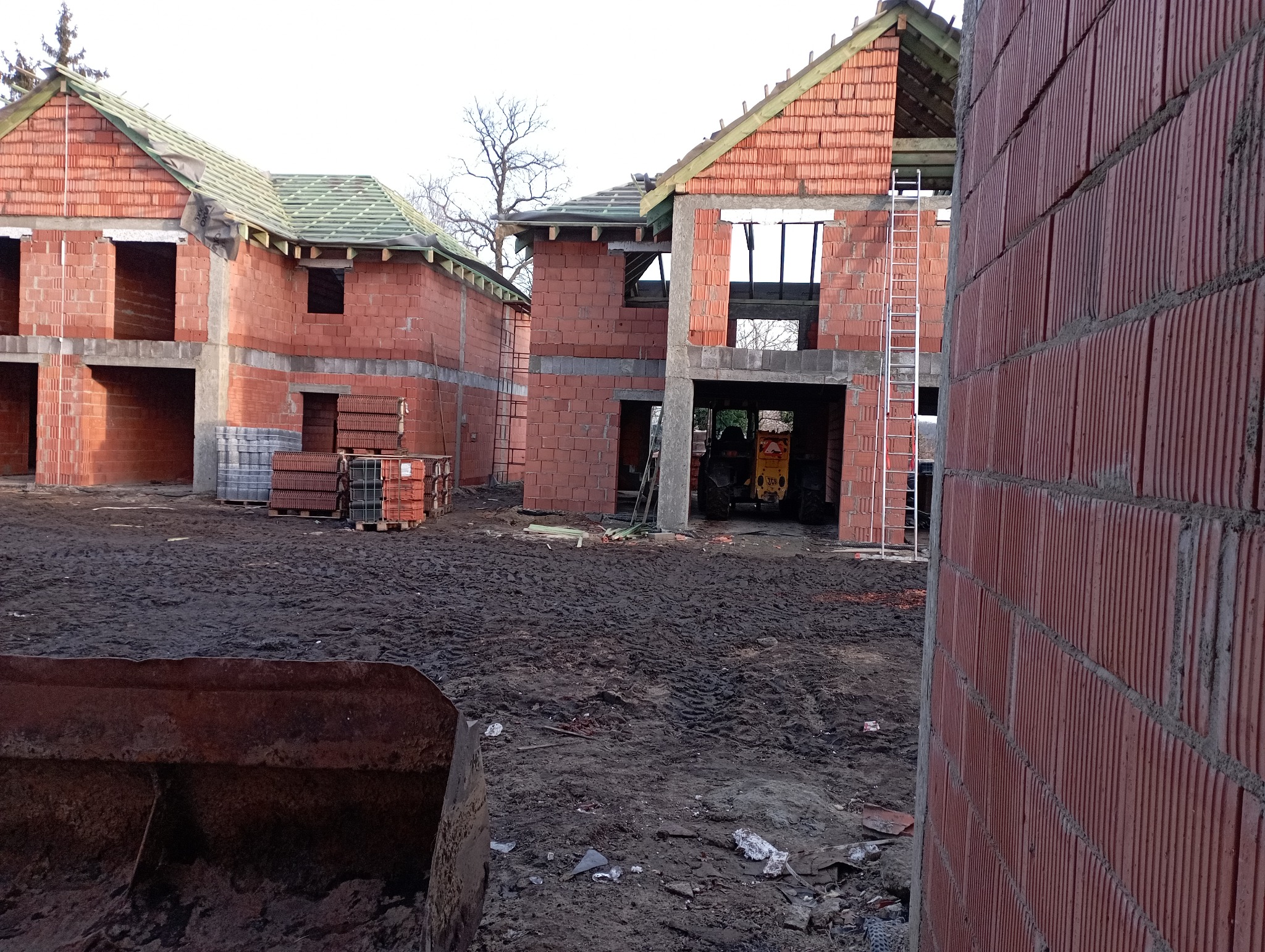 Wznoszenie ścian z czerwonej ceramiki poryzowanej na budowie domów jednorodzinnych, widoczny surowy stan, częściowo zadaszone konstrukcje, teren budowy z błotem, w tle koparka.
