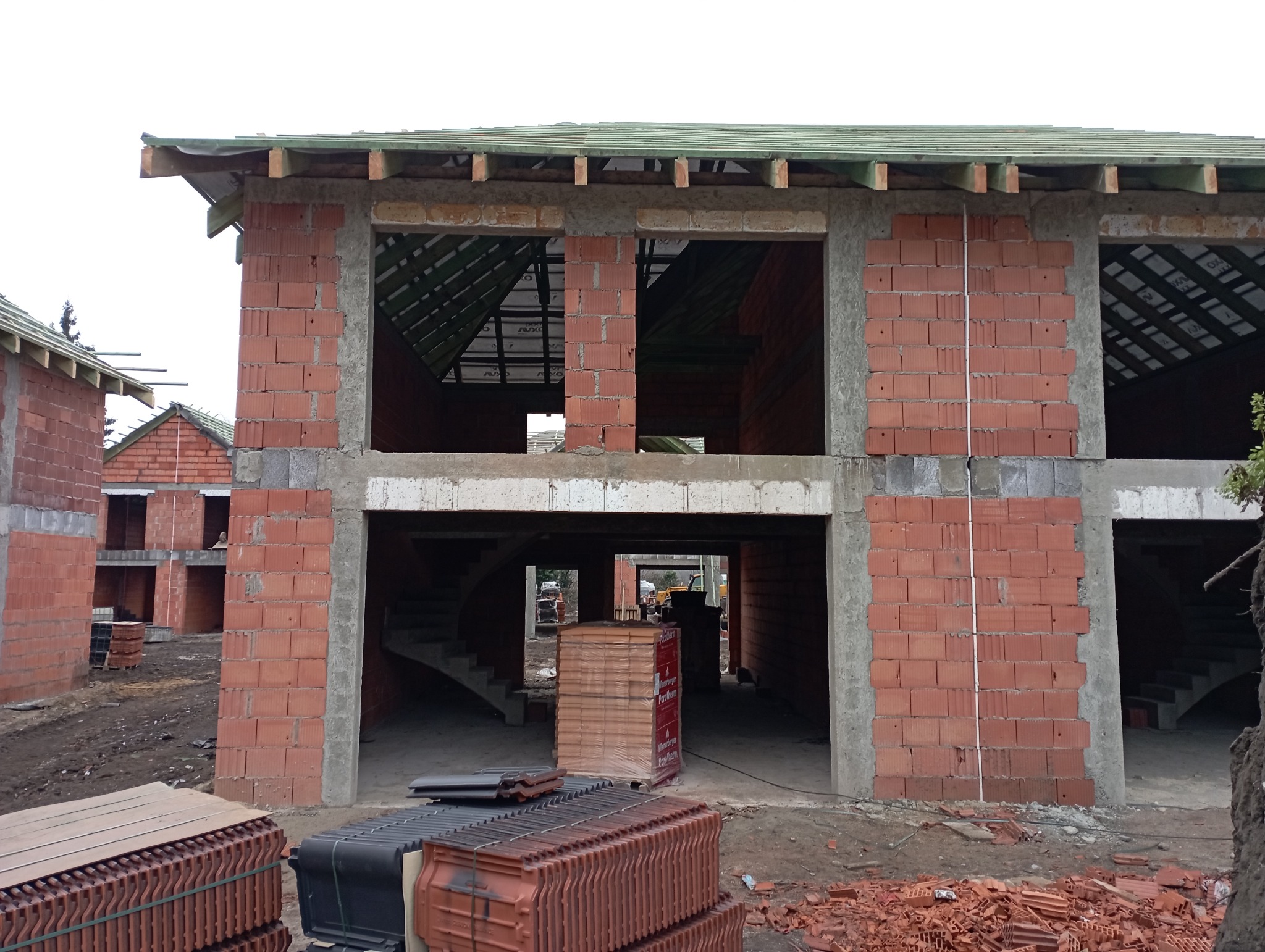Surowy stan domu w trakcie budowy z czerwonej cegły, widoczne puste przestrzenie okienne i drzwiowe, stosy dachówek i cegieł na ziemi, widok na schody wewnętrzne i inne budynki w tle.