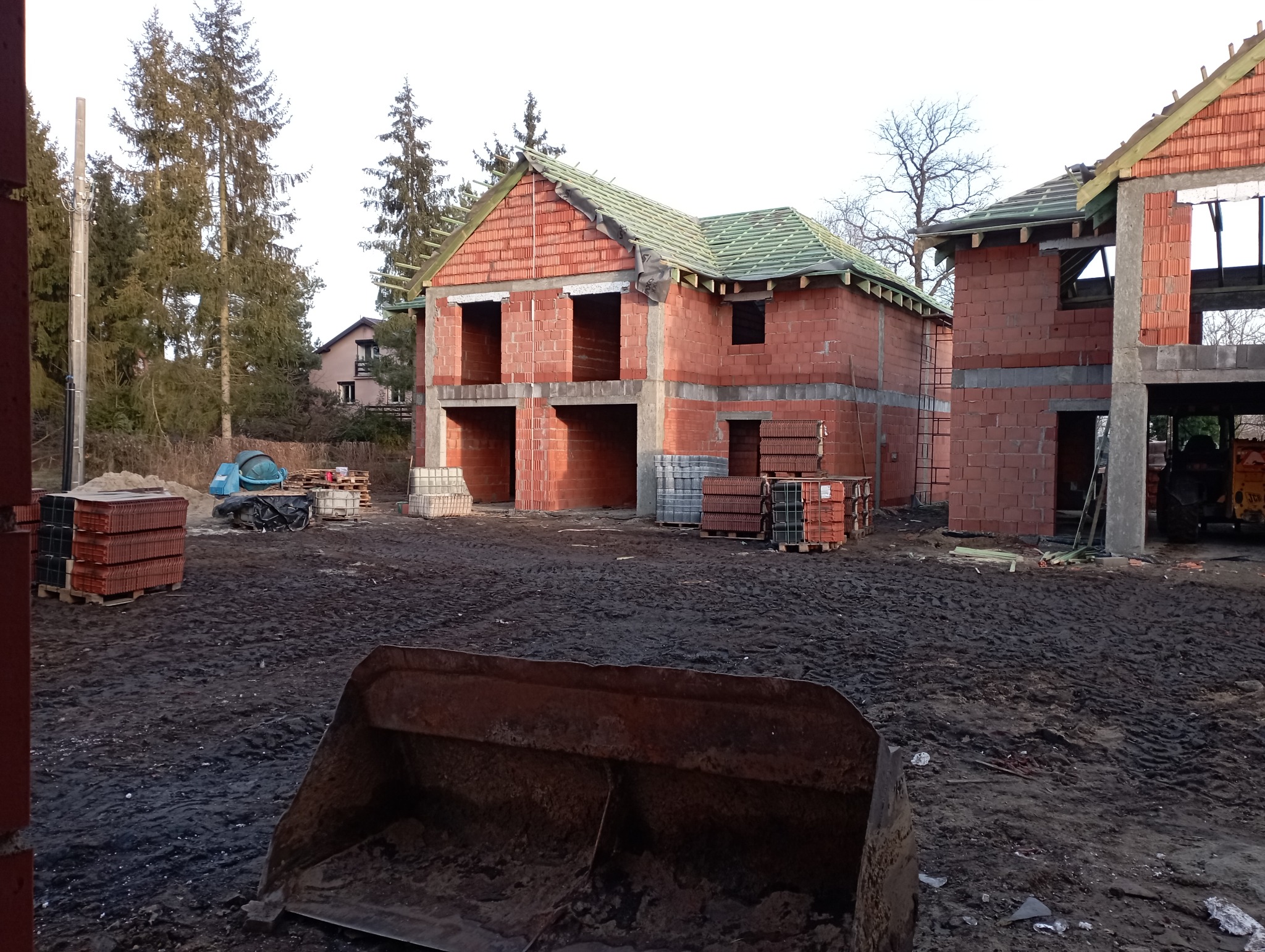 Surowy stan budowy dwóch domów z czerwonej cegły, z widocznymi otworami okiennymi i początkami dachu, na placu budowy z błotnistym podłożem i rozstawionymi materiałami budowlanymi.