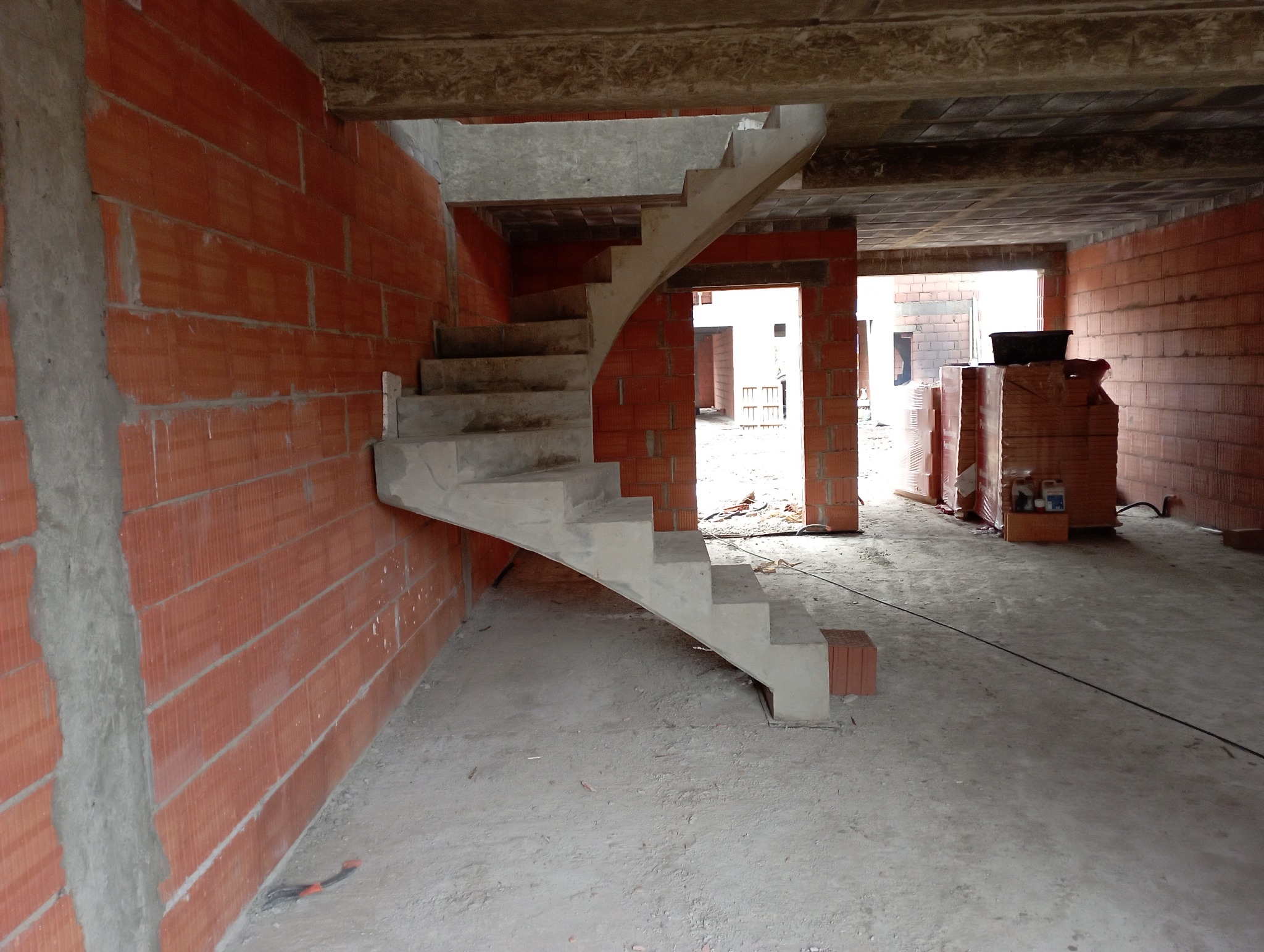 Surowe wnętrze w trakcie budowy: ściany z czerwonej cegły, betonowe schody łukowe prowadzące na wyższą kondygnację, widoczne materiały budowlane i otwory drzwiowe w tle.