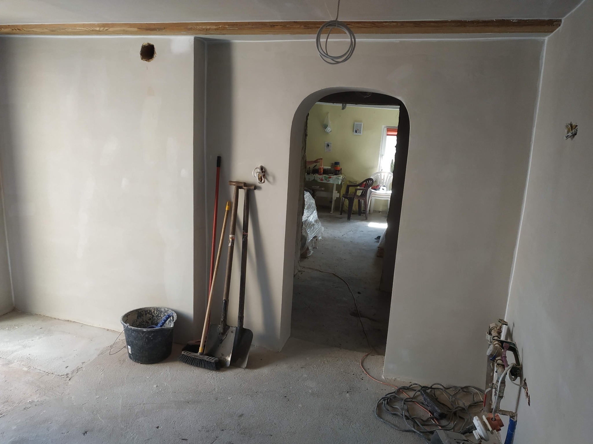 Wnętrze w trakcie prac wykończeniowych: świeżo położone gładzie na ścianach, widoczne narzędzia (szpachle, miotła, wiadro) opierające się o ścianę, surowe drewniane belki pod sufitem, odsłonięte...