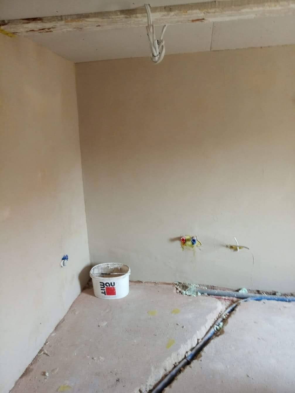 Pokój w trakcie prac wykończeniowych: ściany pokryte świeżą, jasną gładzią, widoczne instalacje elektryczne i hydrauliczne, na podłodze resztki materiałów budowlanych i wiadro po gładzi.