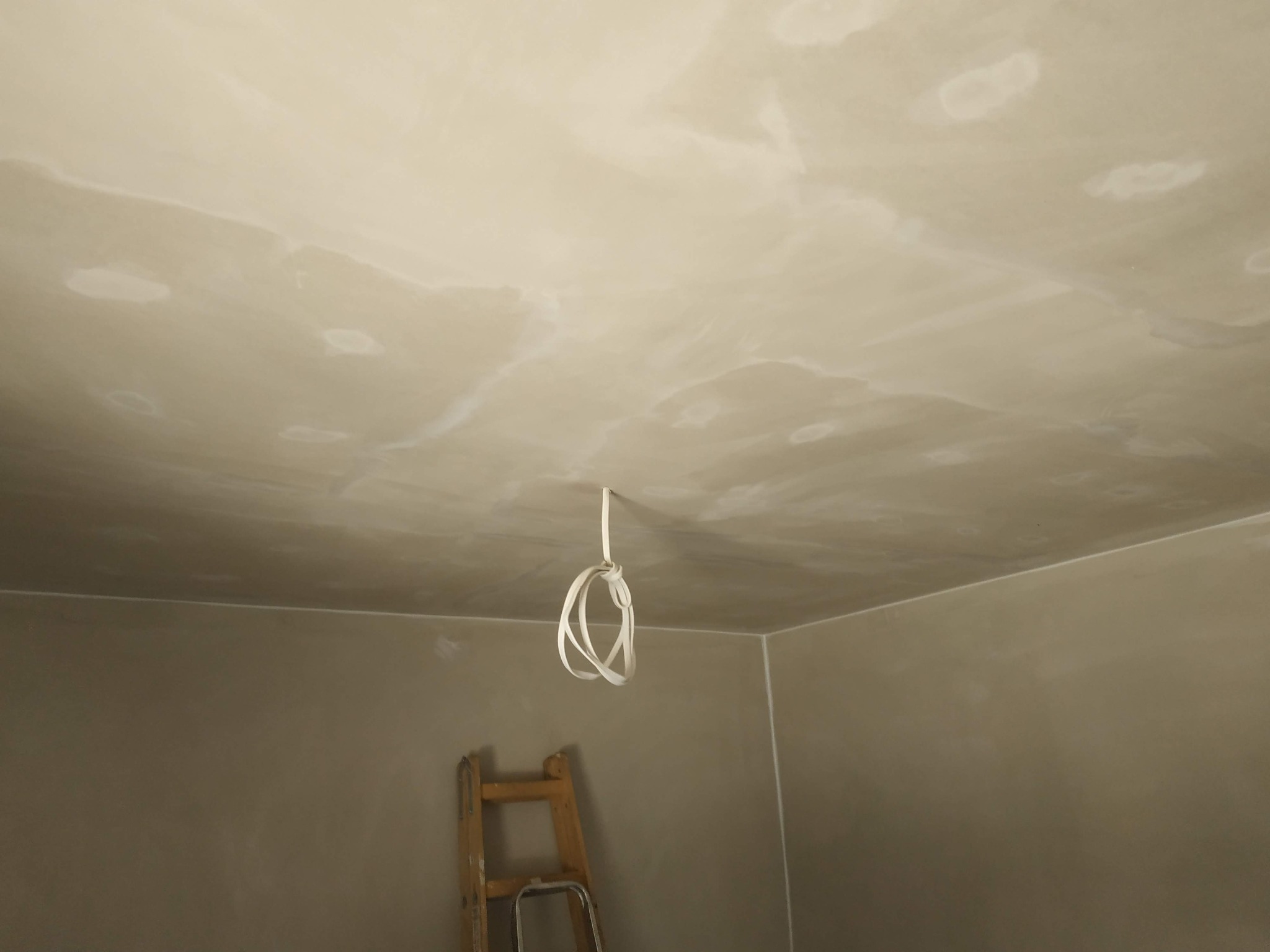 Świeżo nałożona, wilgotna gładź na suficie z widocznymi łączeniami, zwisający kabel elektryczny i fragment drabiny malarskiej w rogu pomieszczenia.