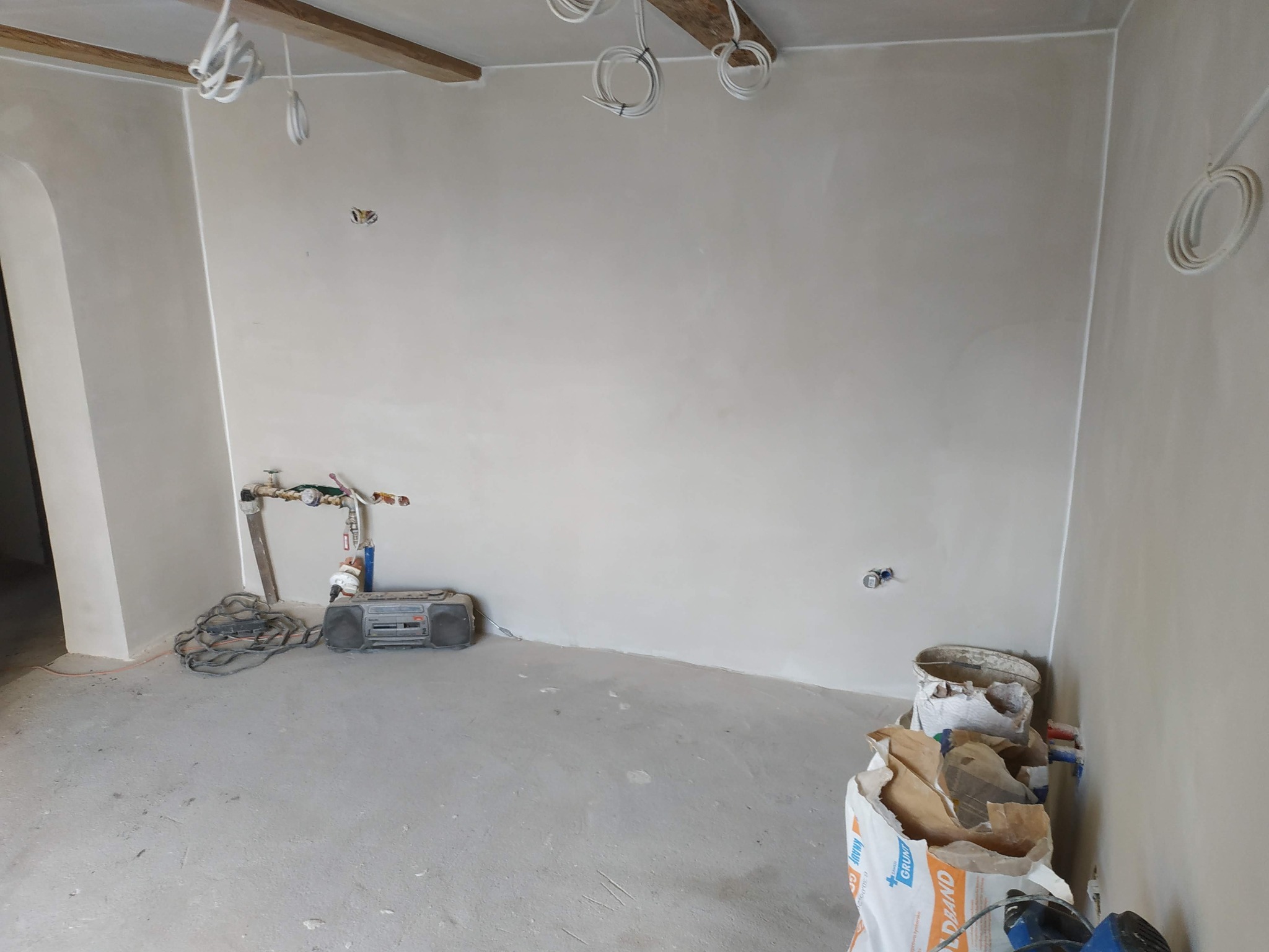 Pokój w trakcie remontu: ściany po nałożeniu gładzi, widoczne instalacje elektryczne, rury wodne, torba z gruntem i radio na podłodze.