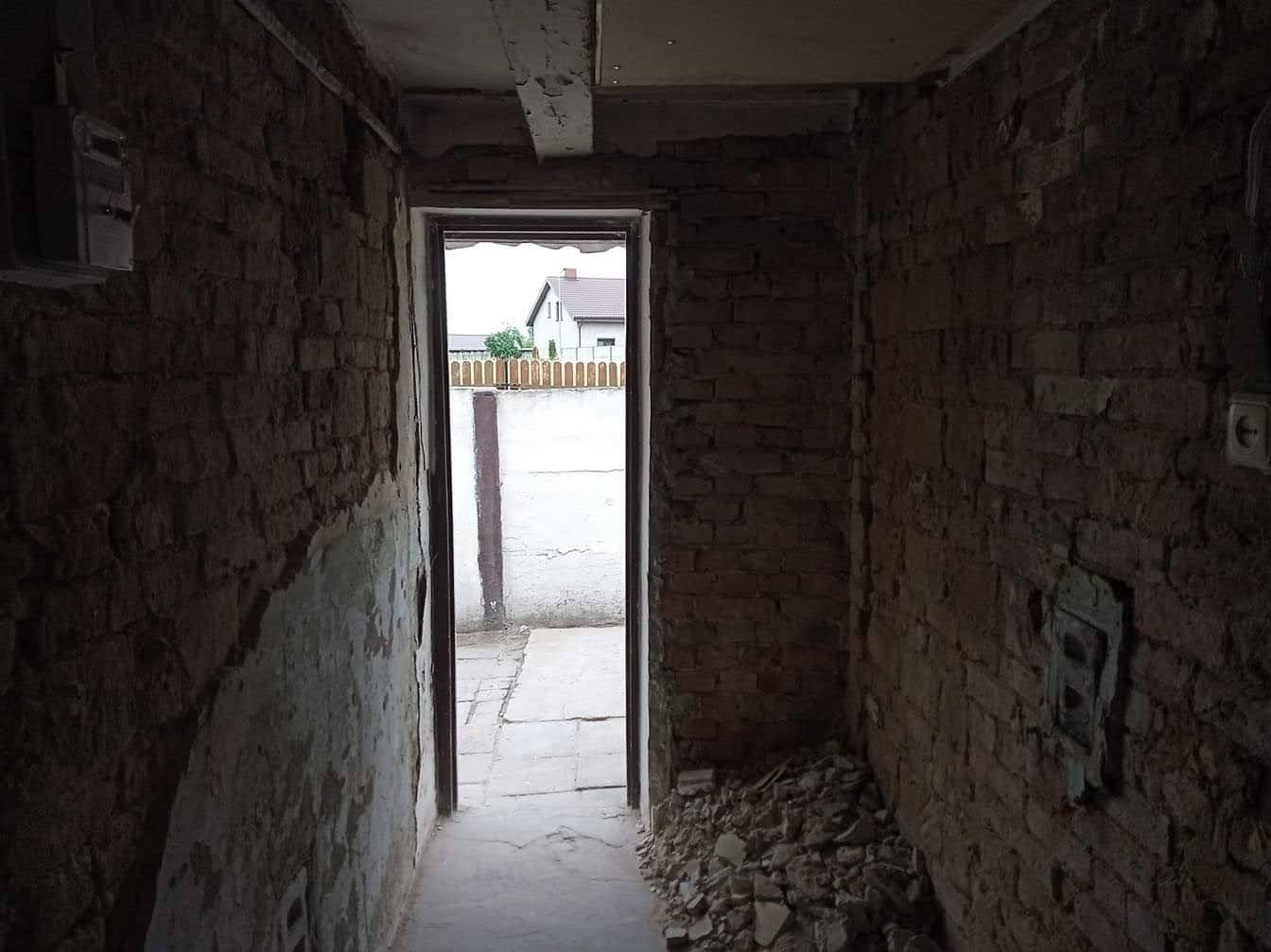 Wnętrze korytarza w stanie surowym z odsłoniętą cegłą, widok na podwórze przez otwarte drzwi, fragmenty starego tynku i gruzu na podłodze.