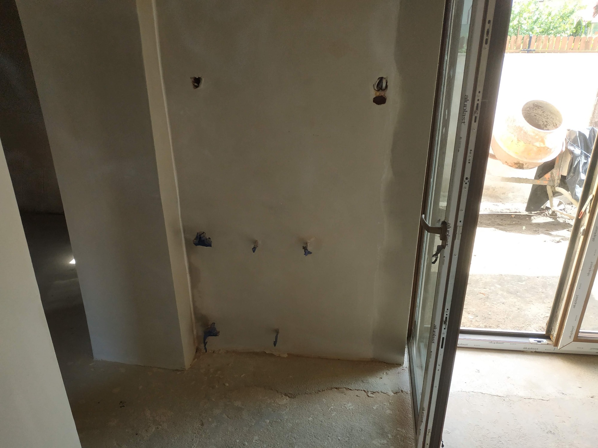 Wnętrze w trakcie prac wykończeniowych: ściana po nałożeniu gładzi z widocznymi otworami na instalacje, częściowo otwarte okno z widoczną betoniarką na zewnątrz.