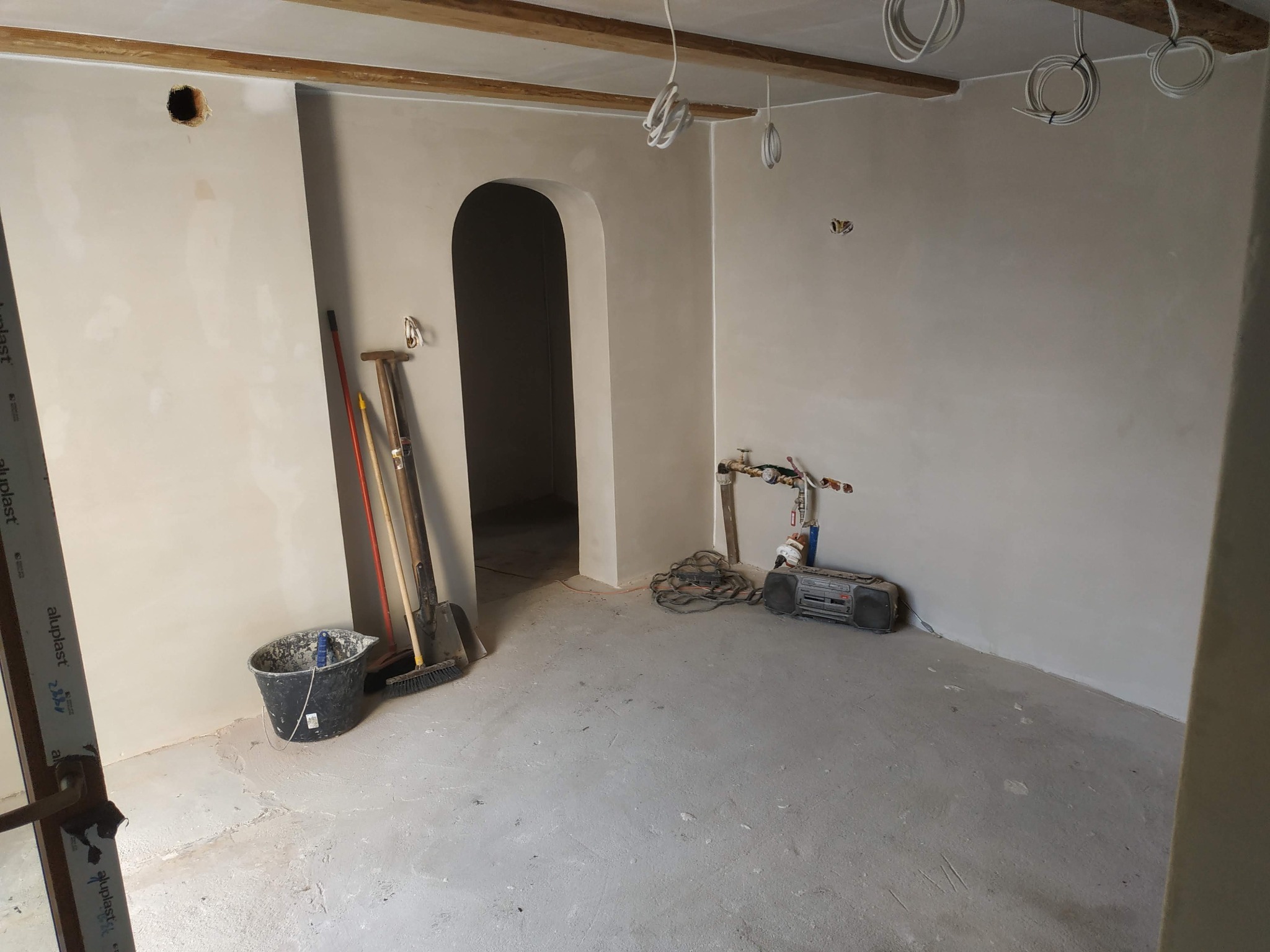 Wnętrze w trakcie prac wykończeniowych: ściany pokryte świeżą warstwą gładzi, widoczne narzędzia malarskie, wiązki kabli zwisające z drewnianego stropu i otwór w ścianie czekający na osprzęt...