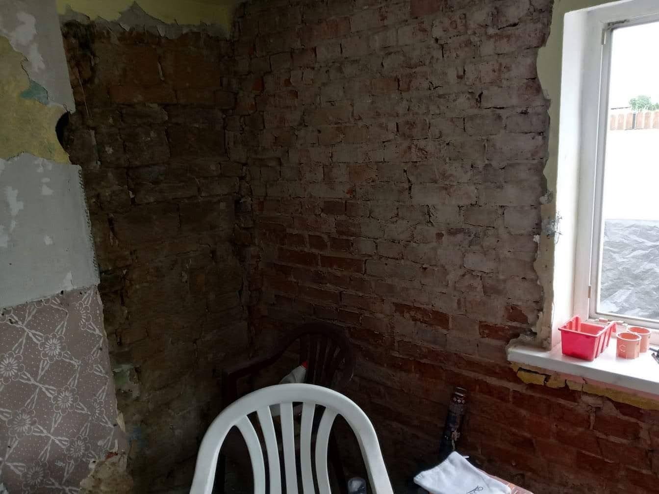 Wnętrze w trakcie remontu: odsłonięta ceglana ściana, fragment okna z parapetem, stare krzesło i plastikowe krzesło na pierwszym planie.