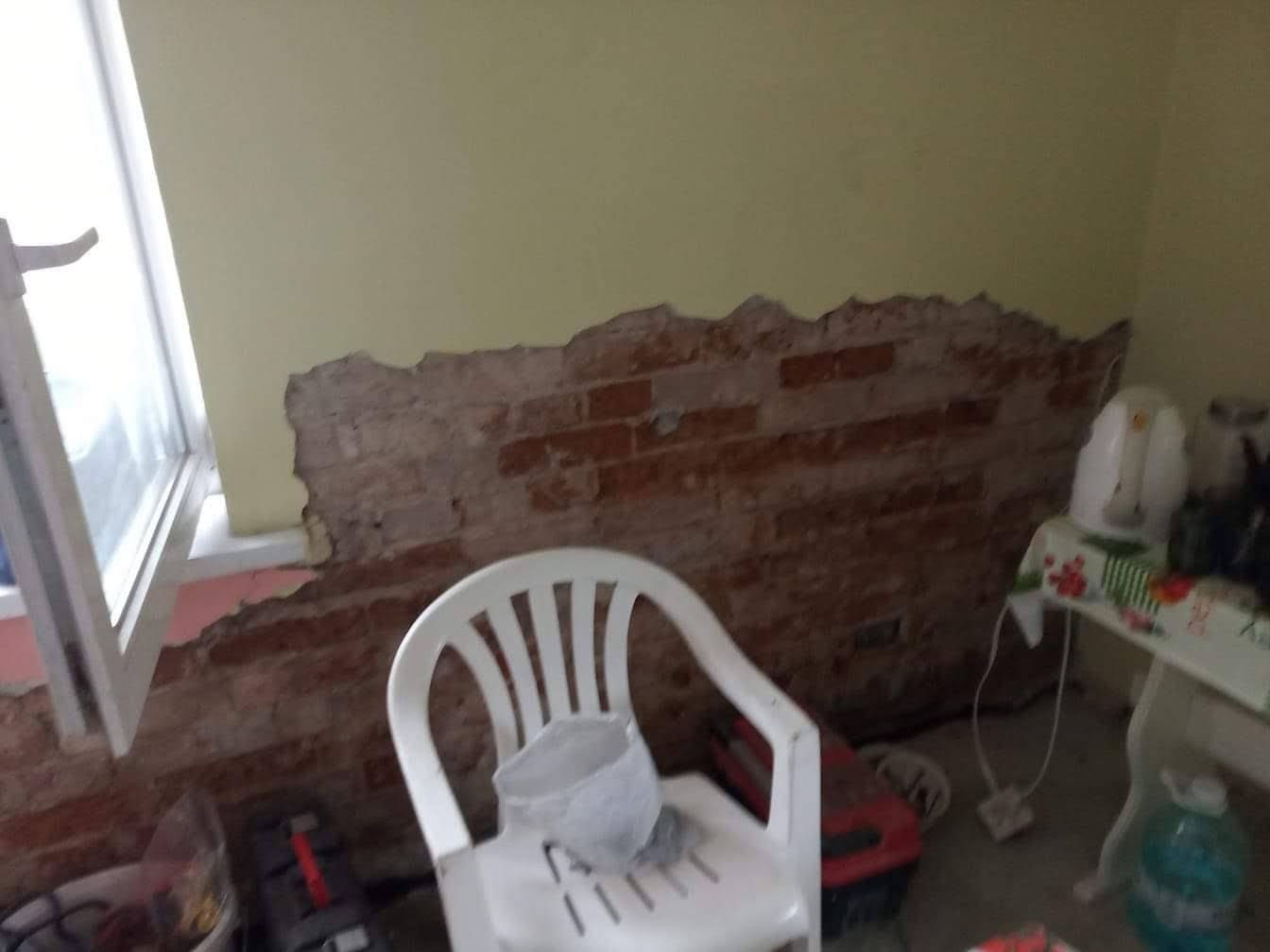Odkryta ceglana ściana w trakcie remontu, z fragmentem otynkowanej ściany powyżej, białe plastikowe krzesło z wiadrem na pierwszym planie, widoczne elementy wyposażenia wnętrza.