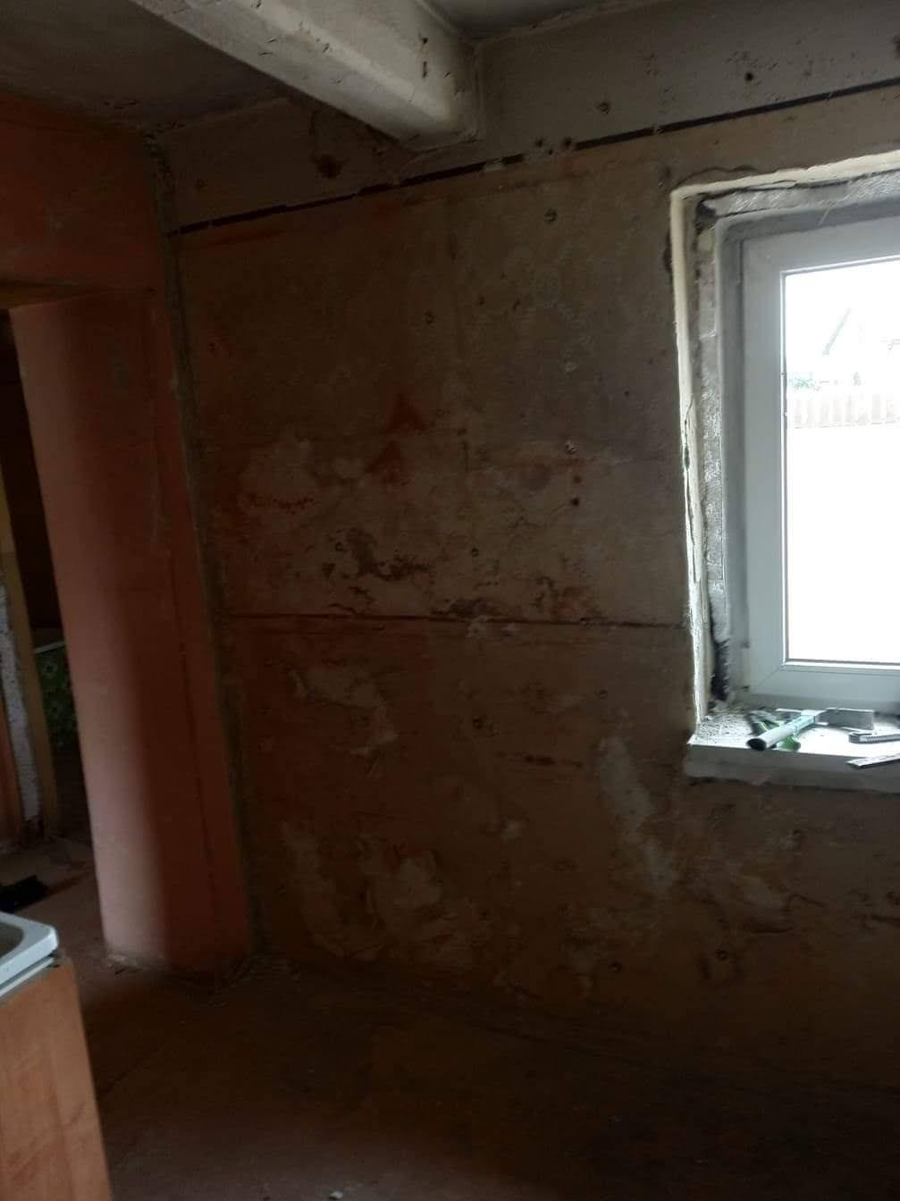 Wnętrze w trakcie prac przygotowawczych do wykończenia, widoczne surowe ściany z pozostałościami farby, okno z narzędziami na parapecie i fragmenty instalacji elektrycznej.
