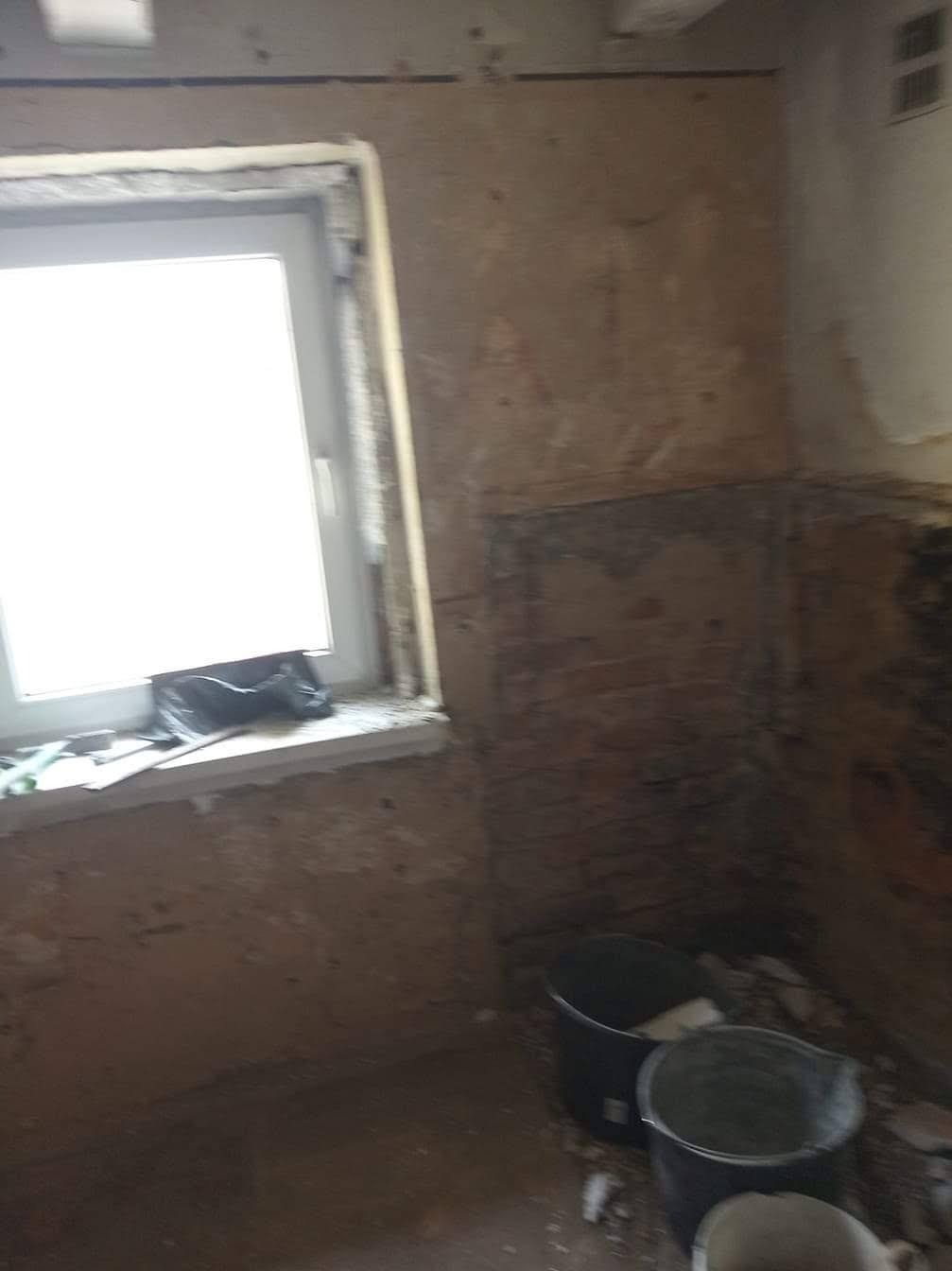 Wnętrze w trakcie prac przygotowawczych do remontu: ściany częściowo odsłonięte z resztek starej okładziny, widoczne fragmenty cegieł, plastikowe wiadra na podłodze, okno z folią ochronną...