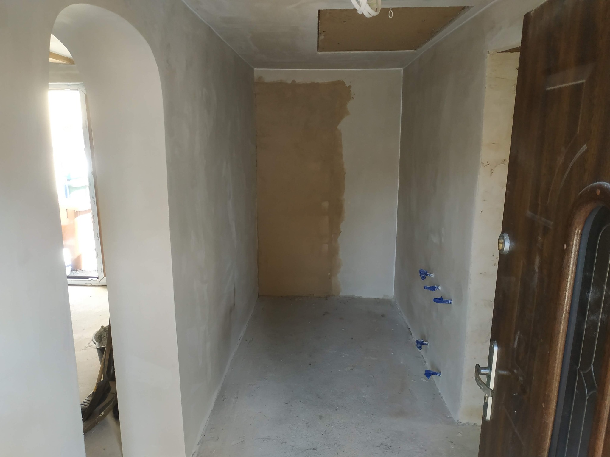 Wnętrze w trakcie remontu: ściany pokryte gładzią, fragment ściany z pozostałościami po starej farbie, niebieskie znaczniki na ścianie, widoczna futryna łukowa i fragment drzwi wejściowych.