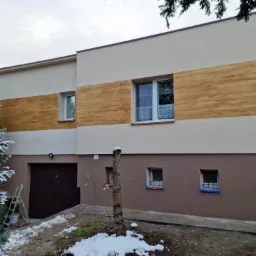Dwukondygnacyjny dom po renowacji elewacji z imitacją drewna na górnej części ściany, widoczna drabina i choinka z resztkami śniegu.