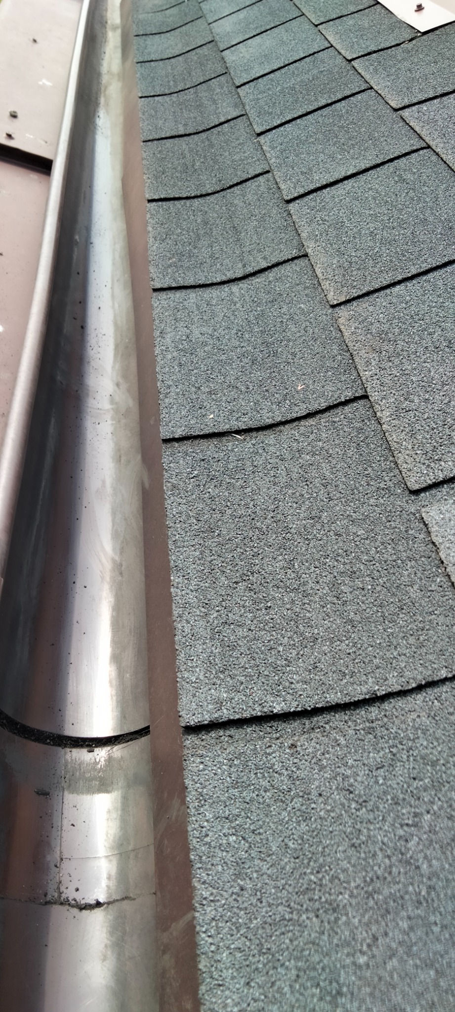 Metalowa rynna, częściowo zabrudzona, przylegająca do dachu pokrytego szarym gontem bitumicznym.