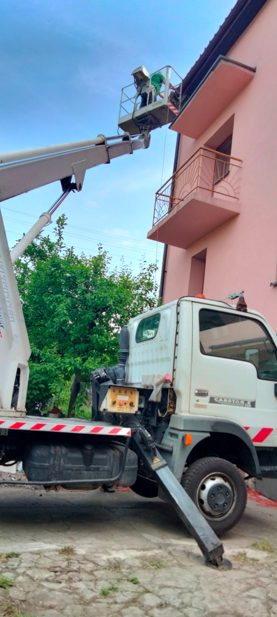 Pracownik w koszu podnośnika teleskopowego, przy różowym budynku z balkonem, wykonuje prace na wysokości.