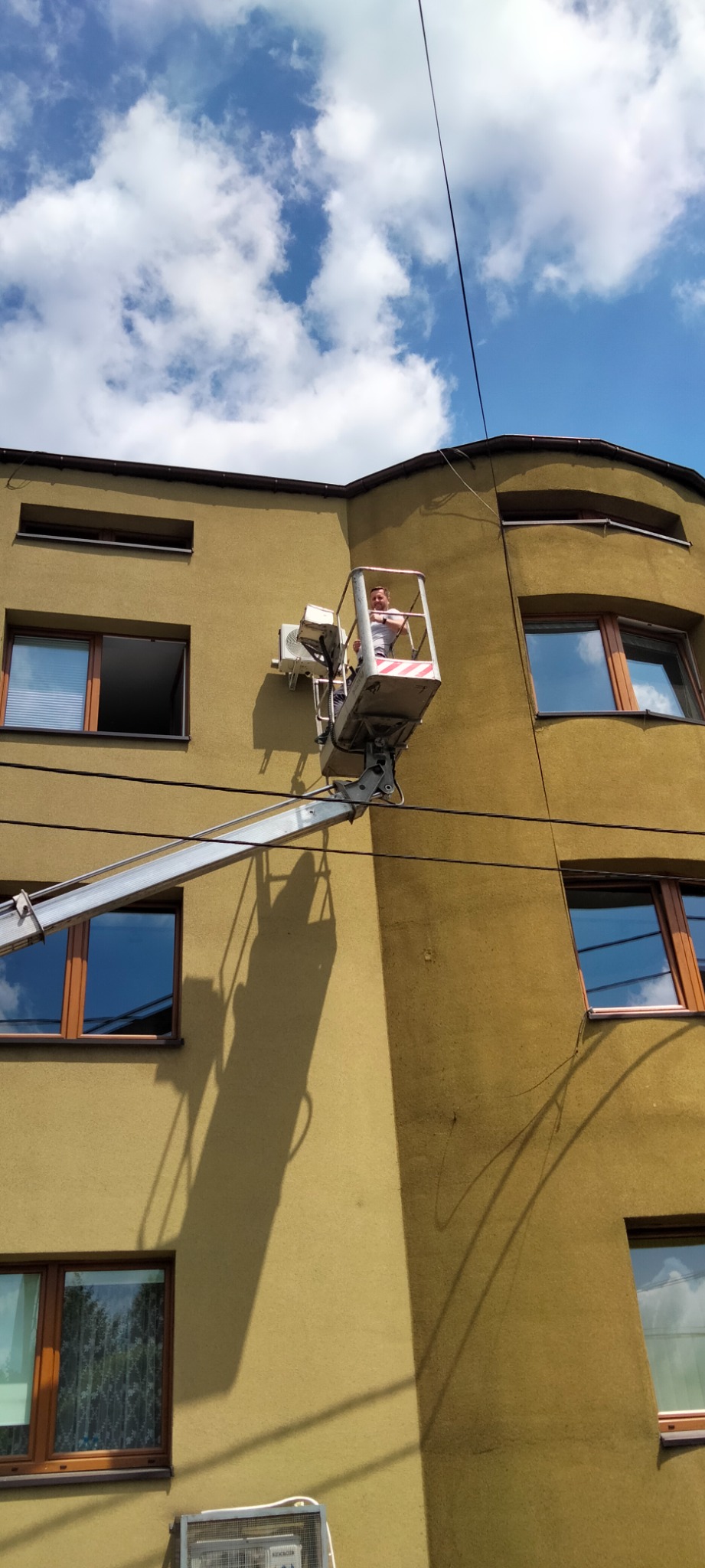 Pracownik na podnośniku koszowym przy ścianie budynku, montujący lub serwisujący klimatyzator zewnętrzny, widok z dołu.