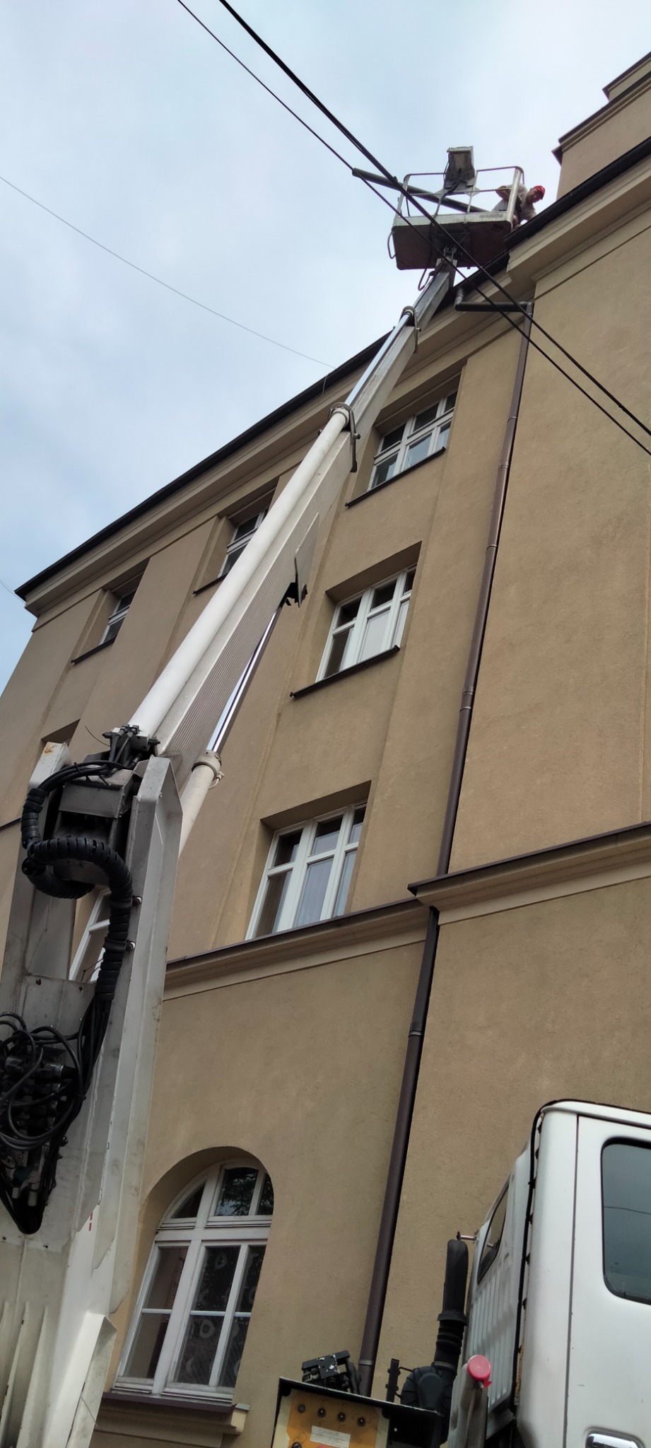 Podnośnik koszowy na tle kamienicy z brązowymi rynnami, pracownik w koszu przy dachu.