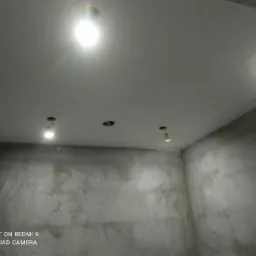 Pokój w trakcie prac wykończeniowych: ściany pokryte szarą warstwą, sufit z zamontowanymi punktami świetlnymi, widoczne otwory na oprawy.