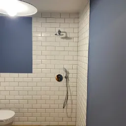 Nowoczesna łazienka z białymi, prostokątnymi płytkami, drewnopodobną podłogą i grafitową ścianą. Widoczna toaleta, deszczownica i bateria podtynkowa. Minimalistyczna lampa sufitowa.