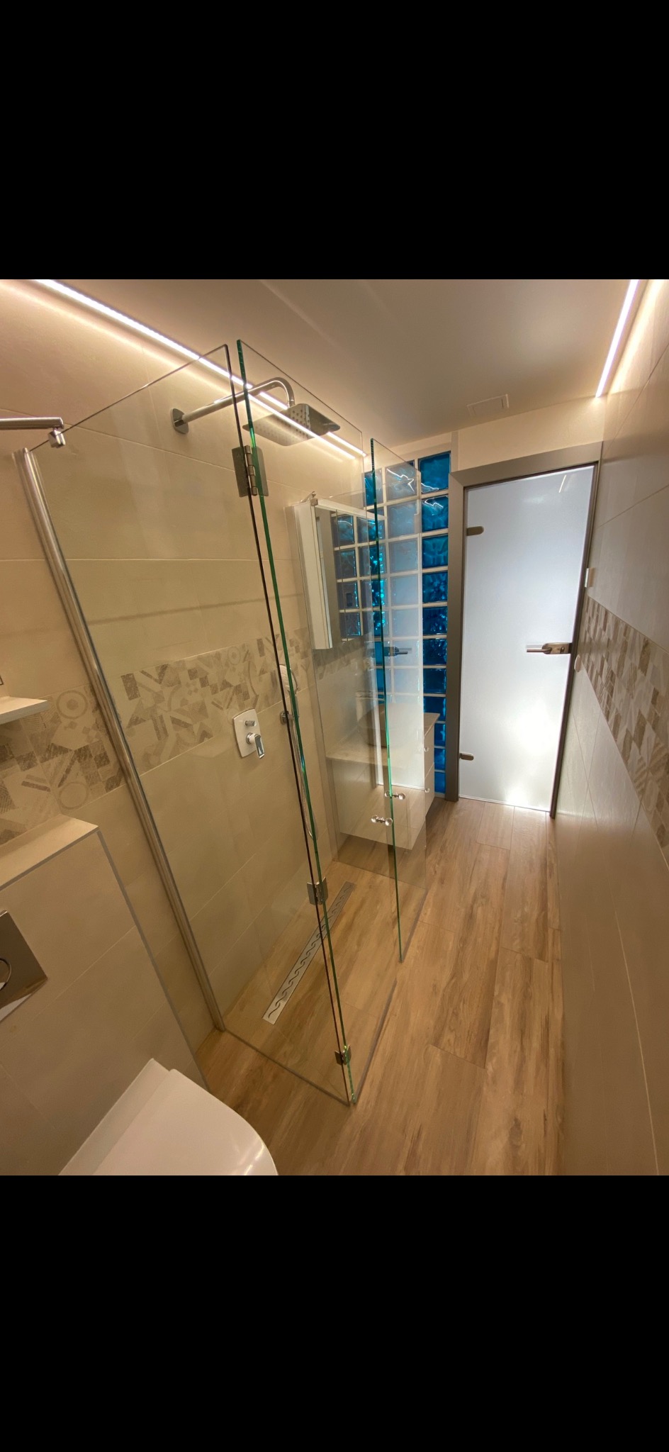 Nowoczesna łazienka z kabiną prysznicową ze szkła hartowanego, drewnopodobną podłogą, ścianami w beżowych odcieniach z dekoracyjnym wzorem i oświetleniem LED podsufitowym.