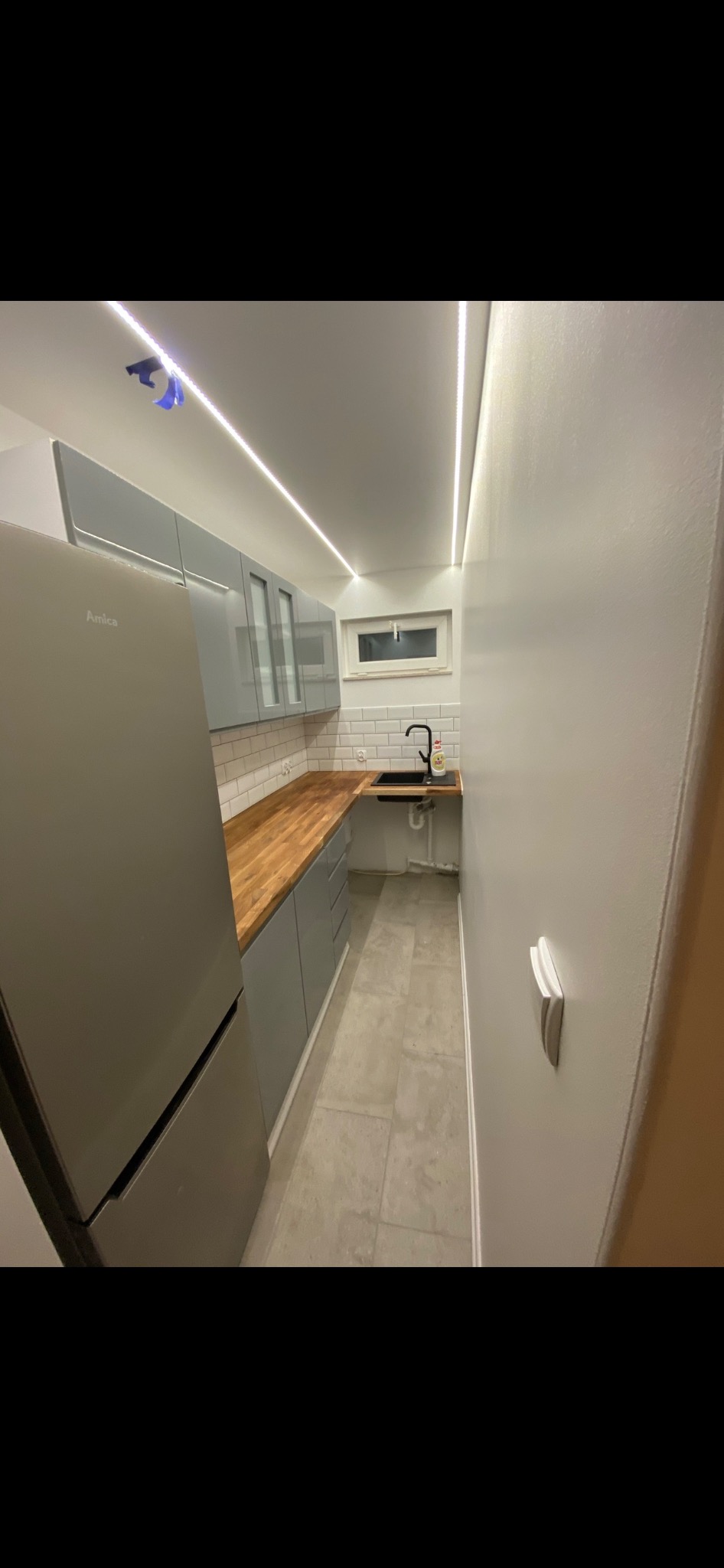 Wąska kuchnia w Bydgoszczy po remoncie, z szarymi szafkami, drewnianym blatem, białymi płytkami na ścianie i podłużnym oświetleniem LED pod sufitem.