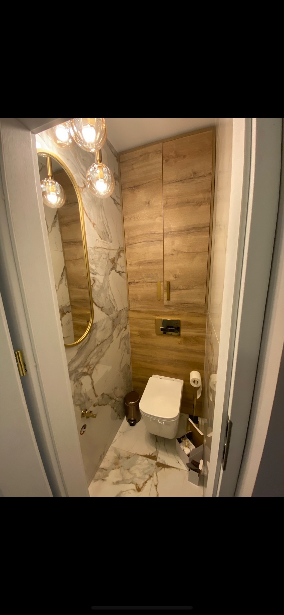 Mała, elegancka toaleta z marmurowymi płytkami, drewnianą szafką, złotymi akcentami i designerską lampą wiszącą z trzema kulami