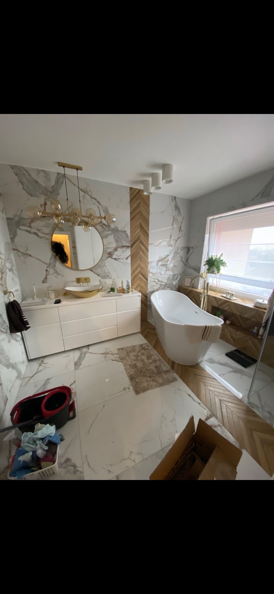 Nowoczesna łazienka z wolnostojącą wanną, marmurowymi płytkami na ścianach i podłodze, oraz drewnianą mozaiką na jednej ze ścian. Widoczna biała szafka z umywalką w kolorze złota i okrągłym lustrem...