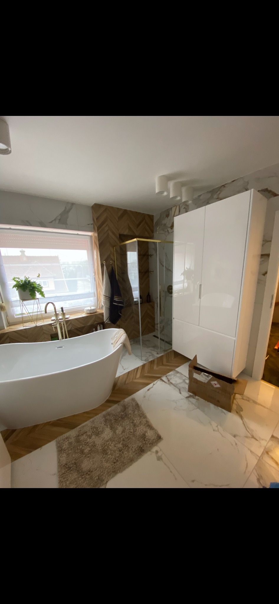 Nowoczesna łazienka z wolnostojącą wanną, kabiną prysznicową ze złotymi ramami, białymi szafkami i marmurowymi płytkami na ścianach i podłodze, wzbogacona o drewniane akcenty i dywanik.