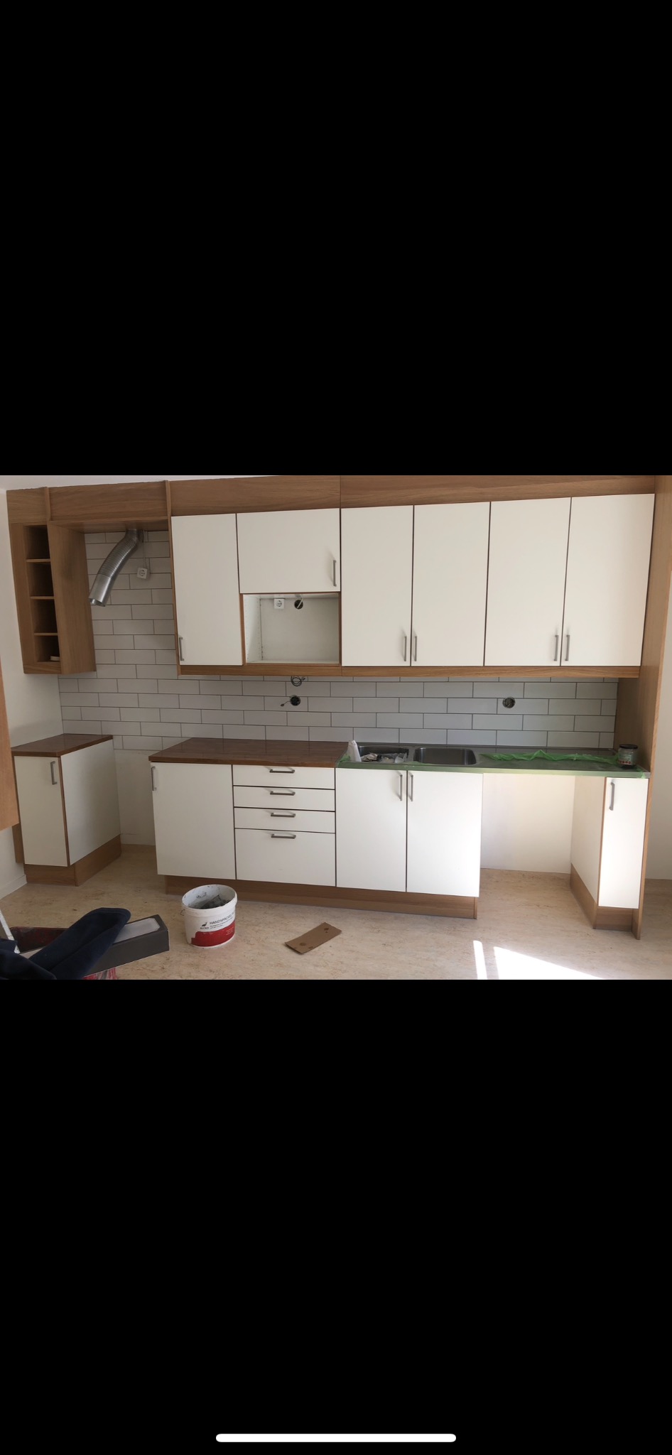 Montaż białej zabudowy kuchennej z drewnianymi akcentami i metalowymi uchwytami, widoczny fragment okładziny z białych płytek i rury wentylacyjnej, na podłodze wiadro z farbą i karton.