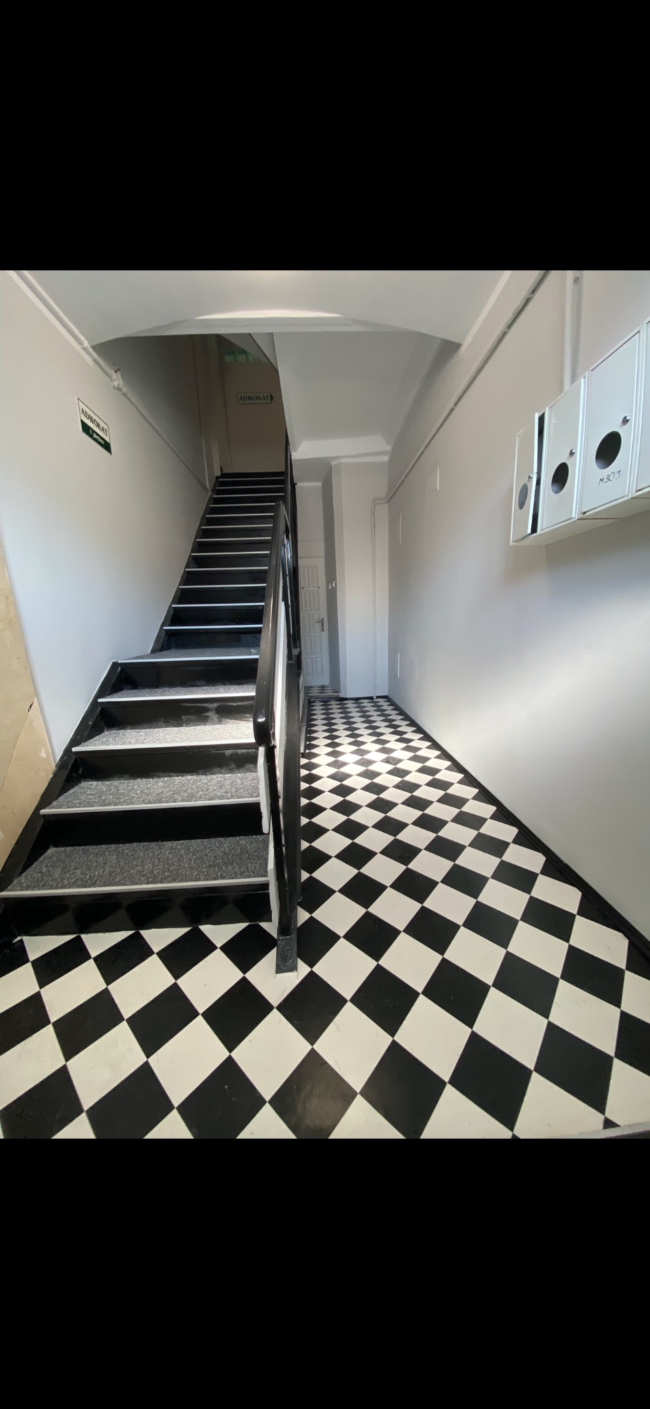 Klatka schodowa z czarno-białą podłogą w szachownicę, szare ściany z tabliczkami 'Adwokat' i białe skrzynki pocztowe z numerami M303.