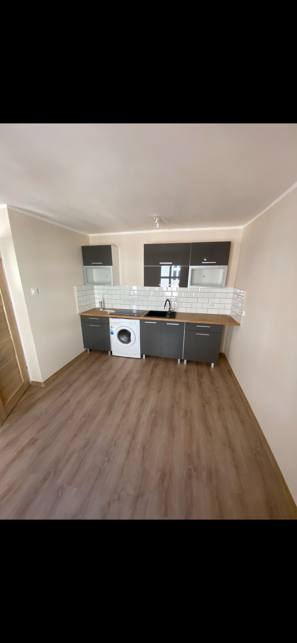 Szare szafki kuchenne z drewnianym blatem i białymi płytkami na ścianie, pralka wbudowana w zabudowę, podłoga z paneli imitujących drewno.