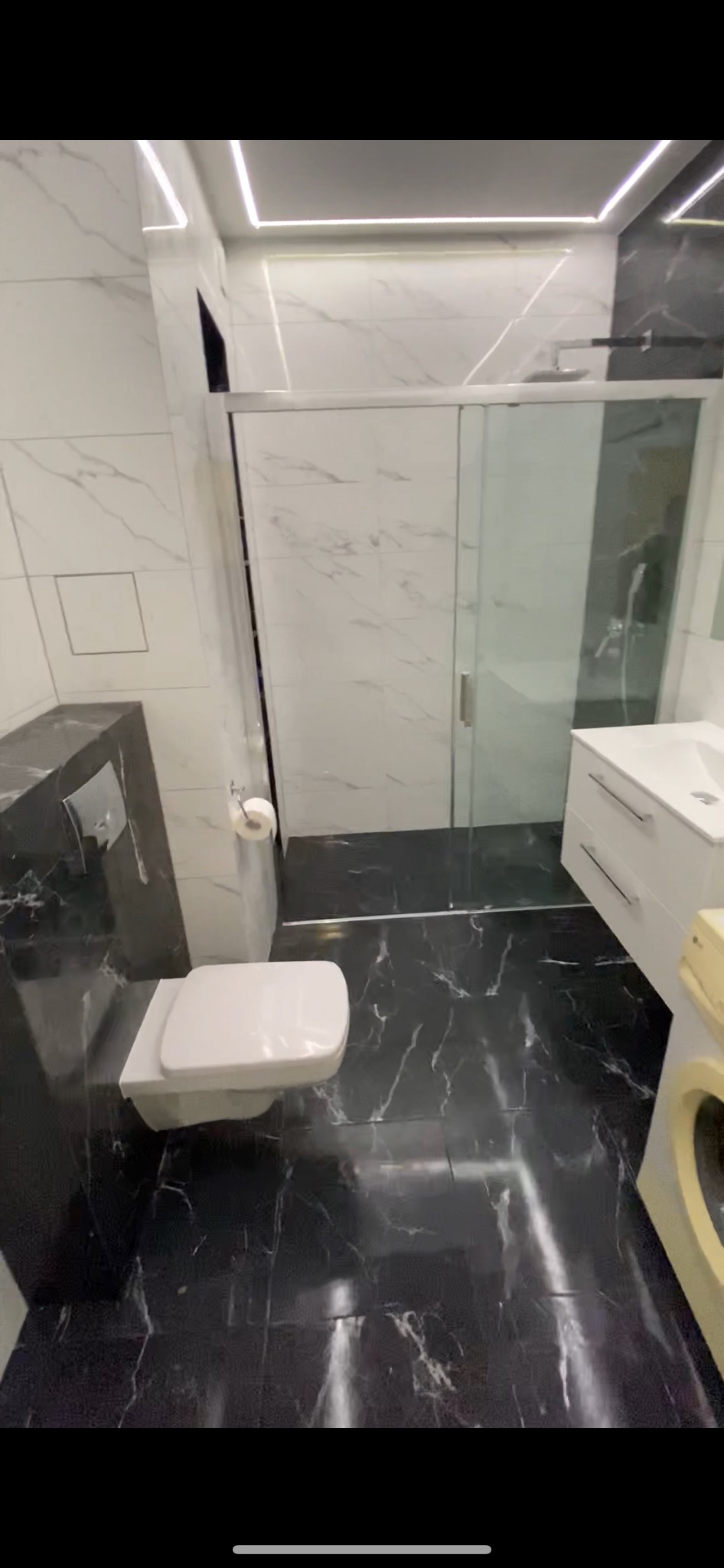 Nowoczesna łazienka z czarno-białymi marmurkowymi płytkami, kabiną prysznicową ze szklanymi drzwiami, podwieszaną toaletą i umywalką z szafką w kolorze białym, oświetleniem LED w suficie.