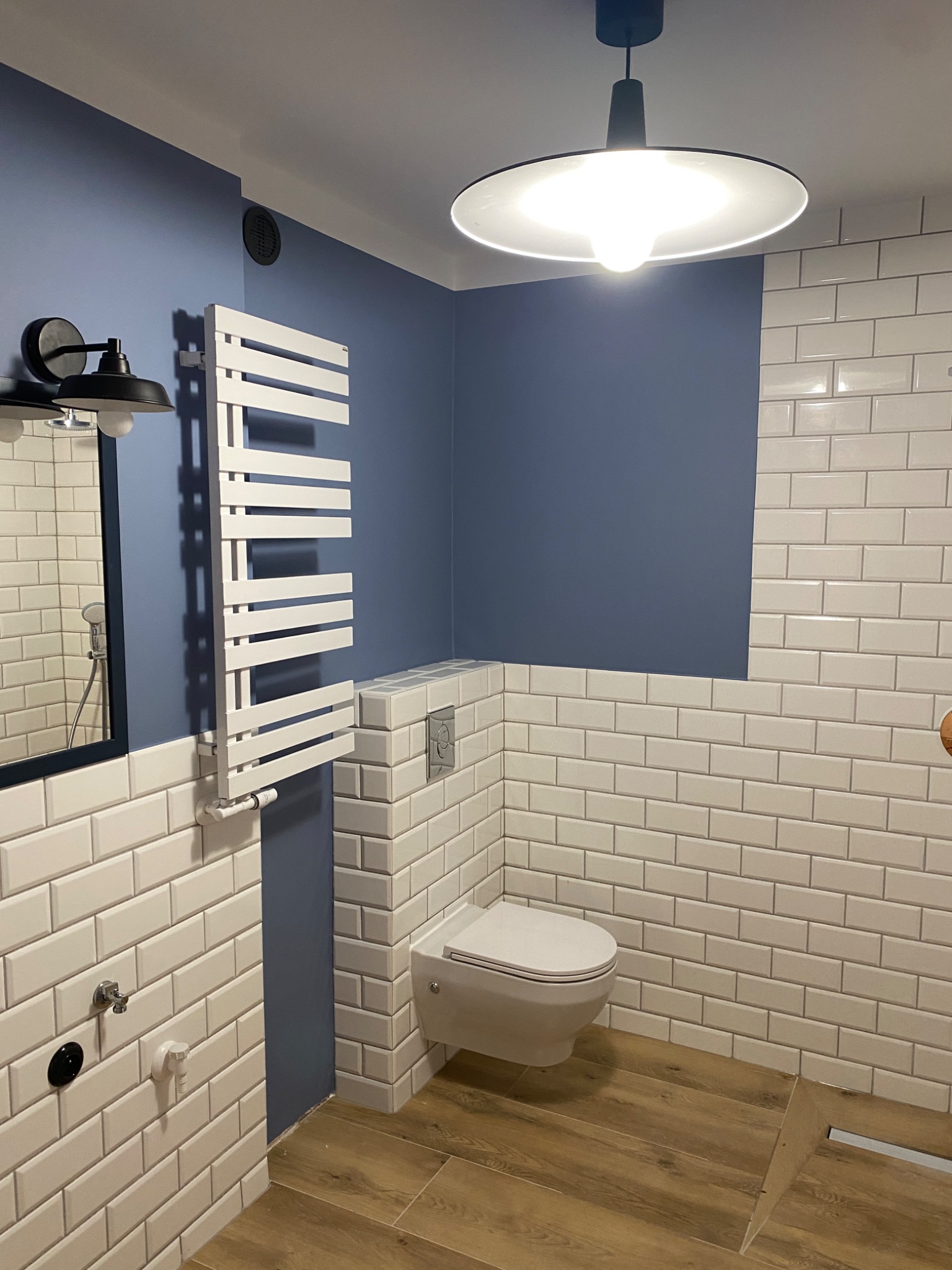 Nowoczesna łazienka z niebieską ścianą, białymi płytkami typu metro i drewnopodobną podłogą, wyposażona w wiszącą toaletę, grzejnik drabinkowy i okrągłą lampę sufitową w stylu industrialnym.