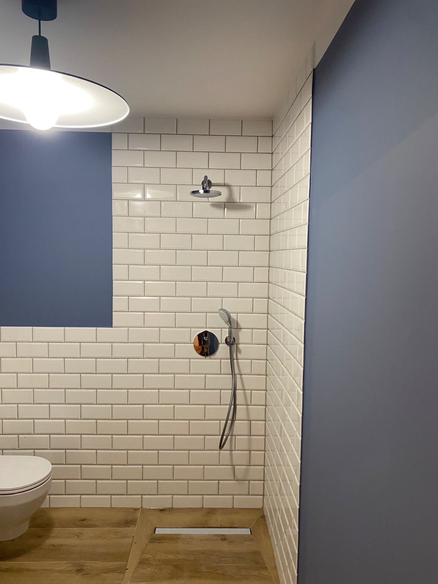 Nowoczesna łazienka z białymi, prostokątnymi płytkami, drewnopodobną podłogą i grafitową ścianą. Widoczna toaleta, deszczownica i bateria podtynkowa. Minimalistyczna lampa sufitowa.