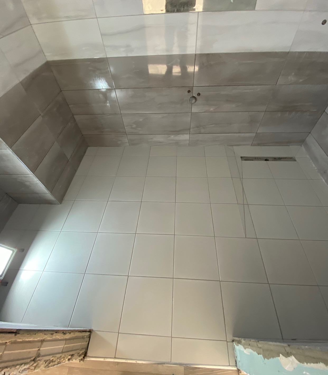 Wnętrze kabiny prysznicowej w trakcie wykańczania, widoczne białe i szare płytki ceramiczne na ścianach i suficie, z zamontowanym odpływem liniowym, częściowo wykończone fugi i niezabezpieczone...