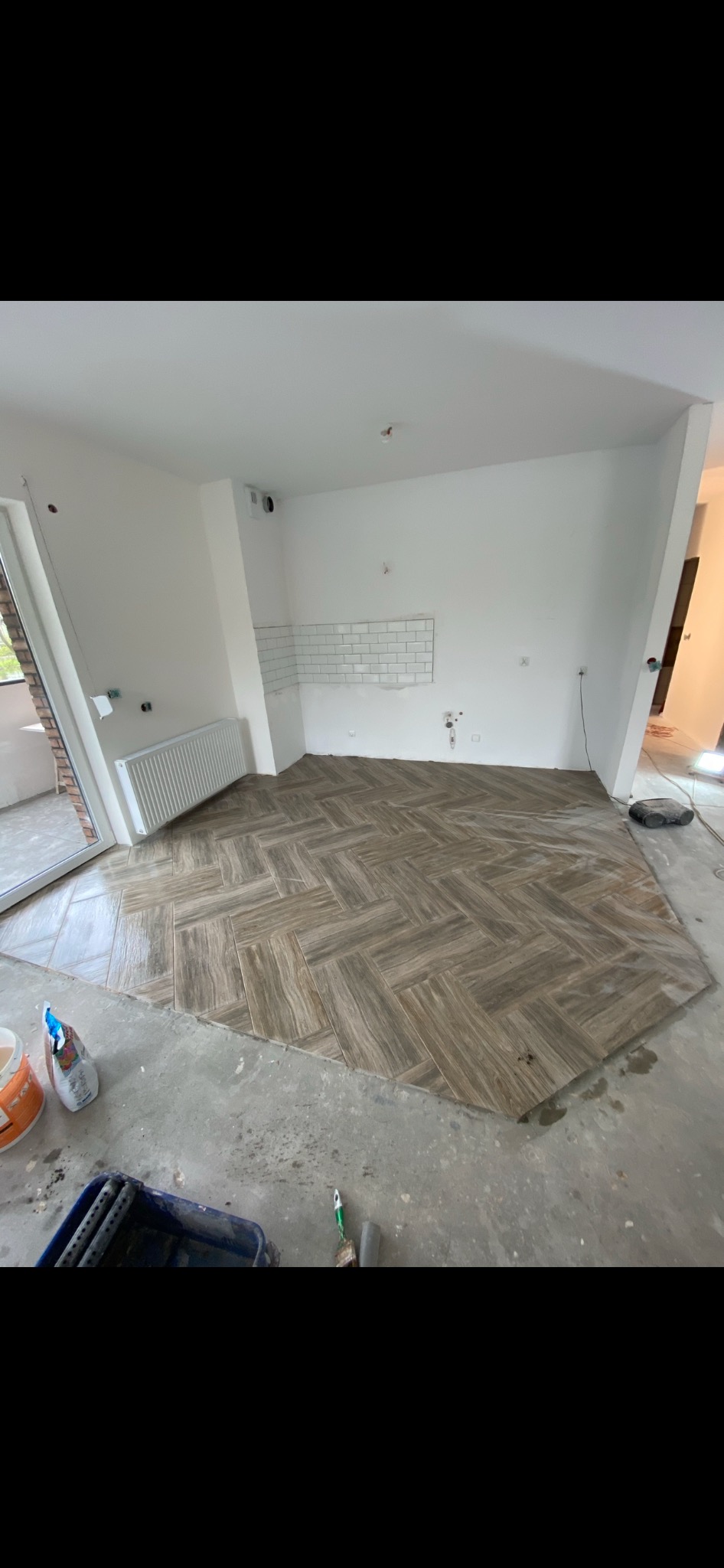 Układanie parkietu w jodełkę z płytek imitujących drewno w trakcie prac wykończeniowych w nowym mieszkaniu, widoczne narzędzia i materiały budowlane, częściowo położona glazura na ścianie w kuchni,...