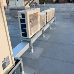 Rząd klimatyzatorów marki Fujitsu zamontowanych na metalowej konstrukcji na płaskim dachu budynku komercyjnego, z widokiem na zalesione wzgórza w oddali.