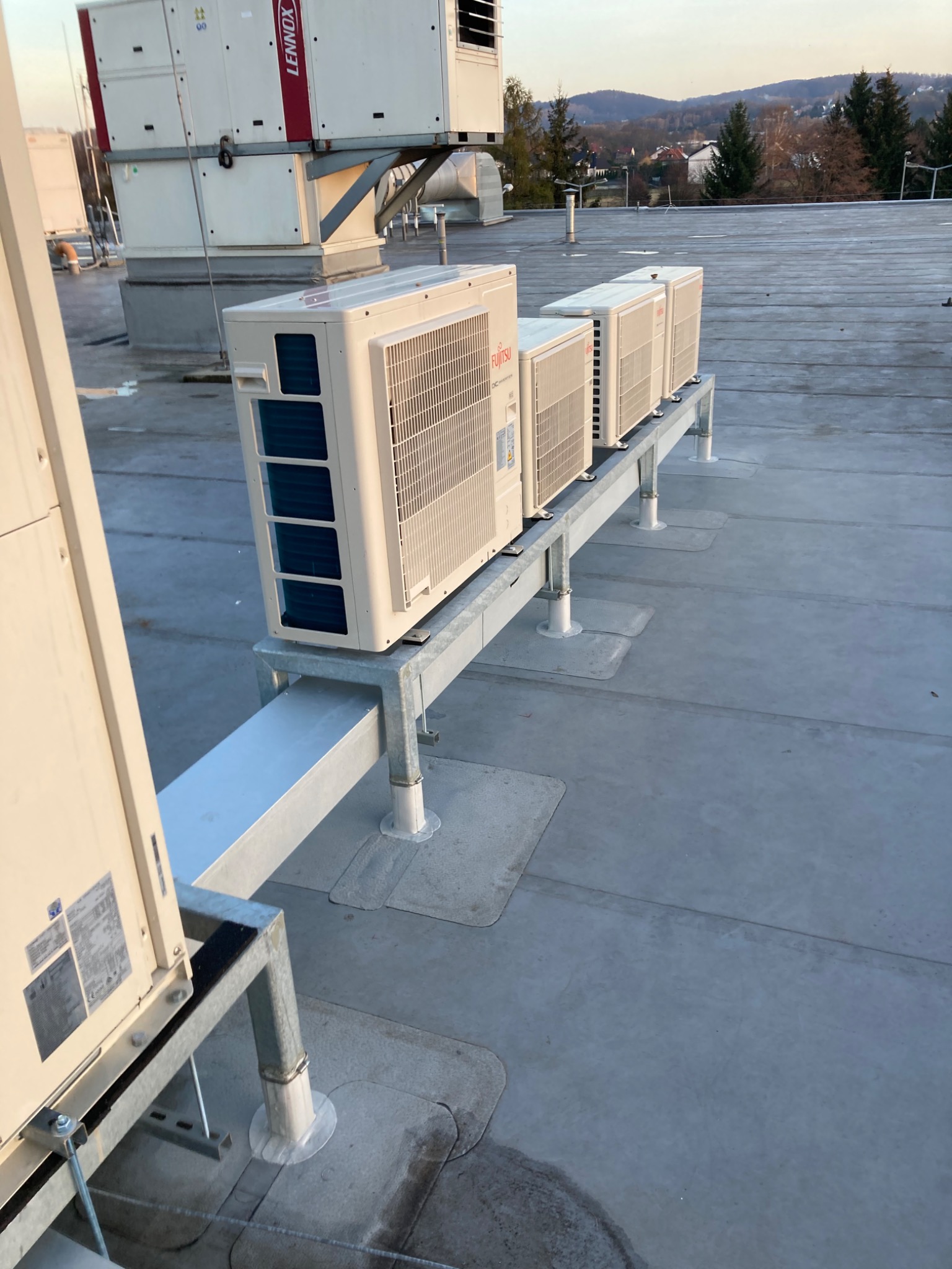 Rząd klimatyzatorów marki Fujitsu zamontowanych na metalowej konstrukcji na płaskim dachu budynku komercyjnego, z widokiem na zalesione wzgórza w oddali.