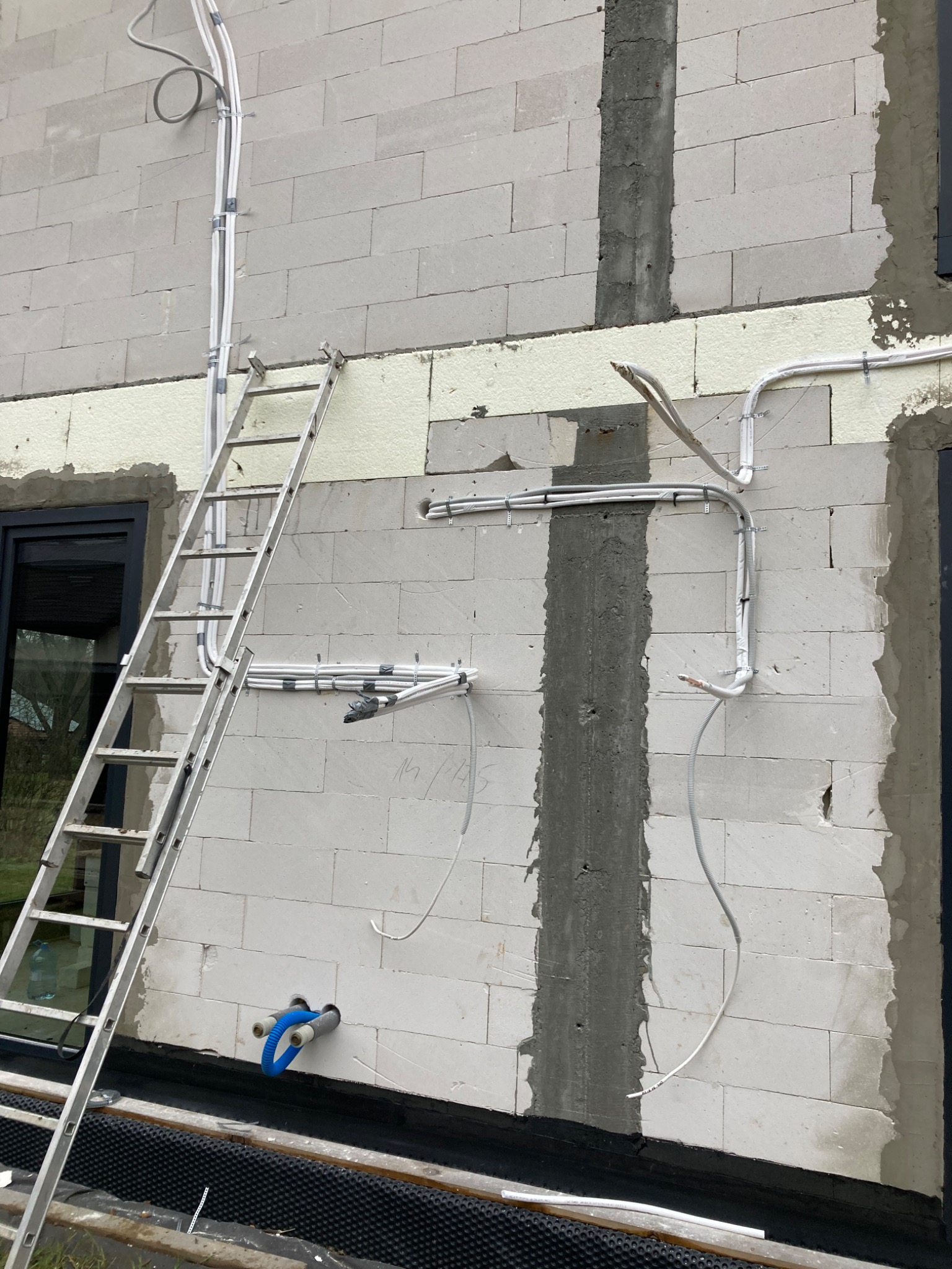 Montaż instalacji wentylacyjnej: widok na ścianę budynku z białych bloczków z widocznymi przewodami wentylacyjnymi, częściowo zamontowanymi, oraz drabina oparta o ścianę.