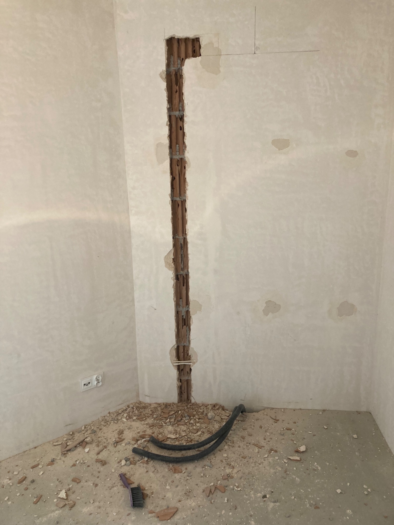 Wykuty pionowy otwór w ścianie z widocznymi rurami, fragment instalacji elektrycznej, gruz na podłodze, pędzelek do zamiatania.