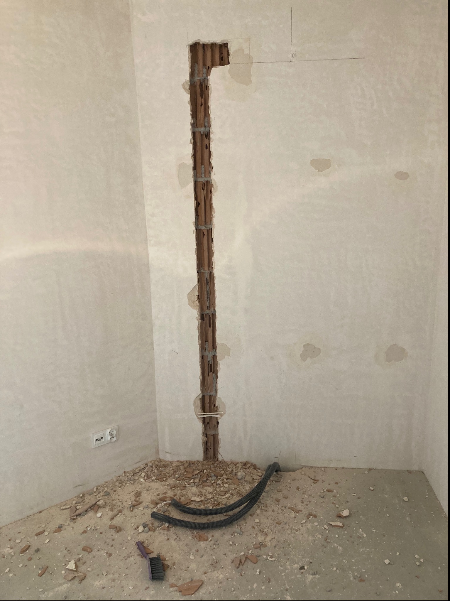 Wykuty w ścianie pionowy kanał instalacyjny z widocznymi rurami, gruzem na podłodze i fragmentem peszla w rogu pomieszczenia.