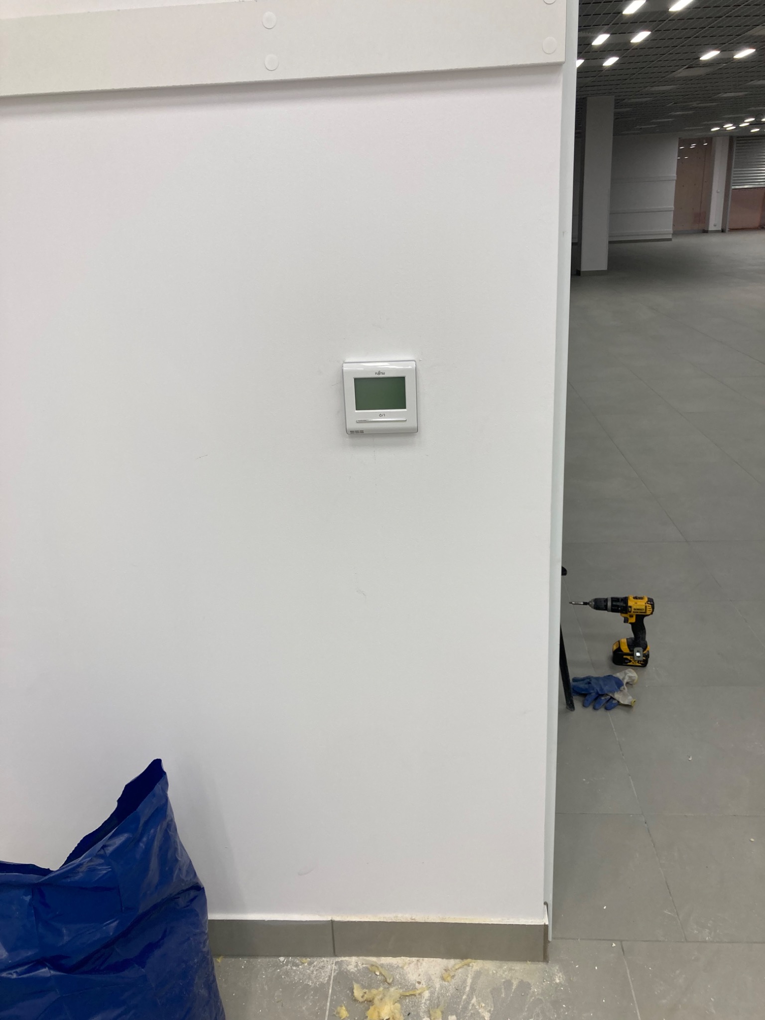 Montaż panelu sterowania klimatyzacją Fujitsu na białej ścianie w budynku komercyjnym, widoczna wiertarka DeWalt i rękawice robocze na szarej podłodze.