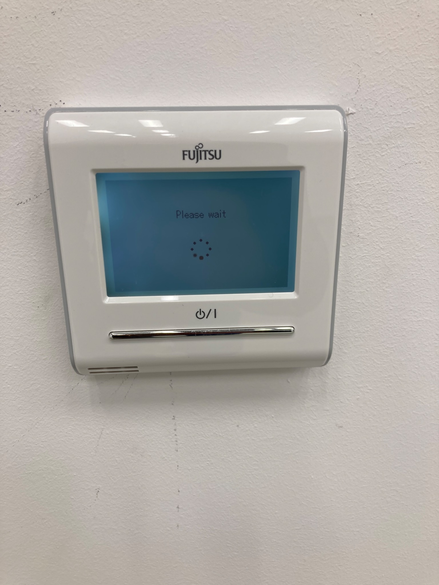 Biały panel sterowania klimatyzacji Fujitsu na białej, lekko zabrudzonej ścianie, wyświetlający komunikat 'Please wait' z obracającym się wskaźnikiem ładowania.