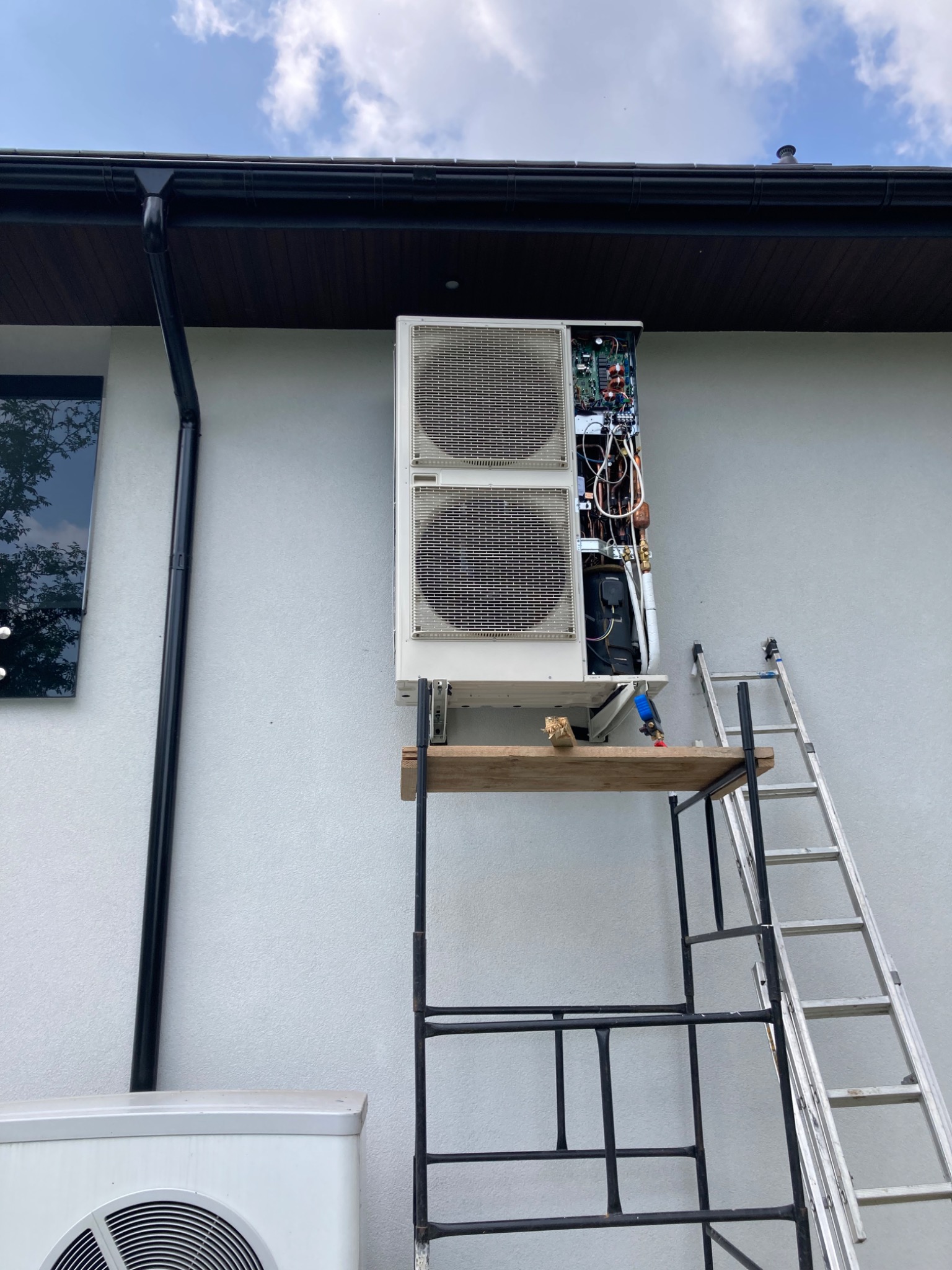 Biała jednostka zewnętrzna pompy ciepła zamontowana na jasnoszarej ścianie budynku, z otwartą obudową ukazującą wewnętrzne komponenty, wsparta na rusztowaniu z drewnianą deską i drabina oparta...