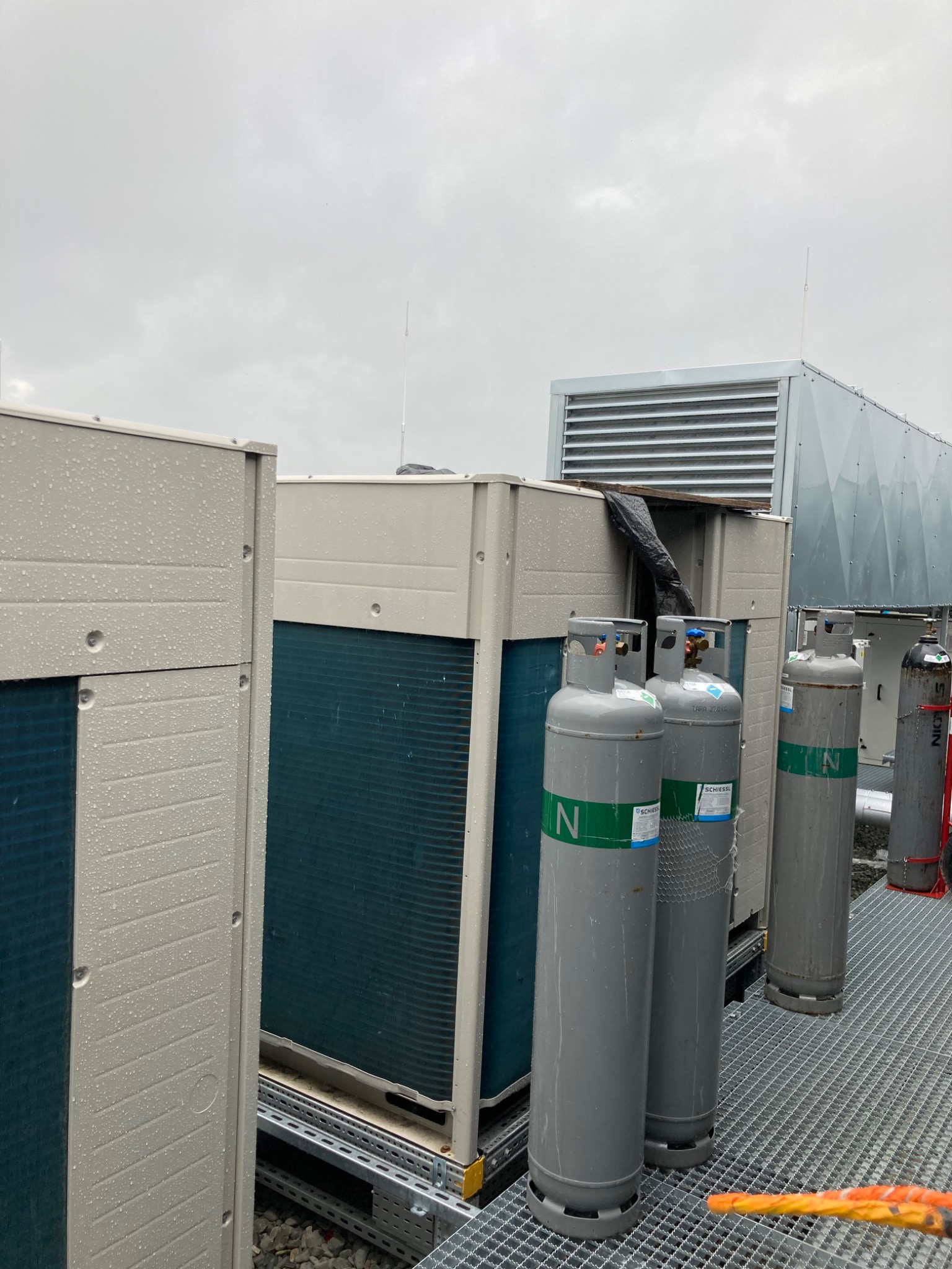 Instalacja klimatyzacji na dachu budynku, widoczne jednostki zewnętrzne i butle z azotem, mokre od deszczu, na metalowej kratownicy.