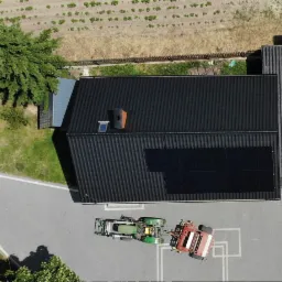 Widok z góry na dom z czarnym dachem, na którym zamontowane są panele fotowoltaiczne; na podwórzu stoi traktor z przyczepą i samochód osobowy.