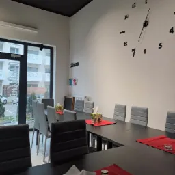 Nowoczesna sala konferencyjna z długim, czarnym stołem, szarymi krzesłami, dekoracyjnym zegarem na ścianie i pudełkami chusteczek na czerwonych podkładkach.