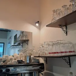 Bar z profesjonalnym ekspresem do kawy, stertą białych filiżanek i talerzyk&oacute;w, oraz p&oacute;łkami z czystymi szklankami i kieliszkami do wina, oświetlony ciepłym światłem żar&oacute;wek.